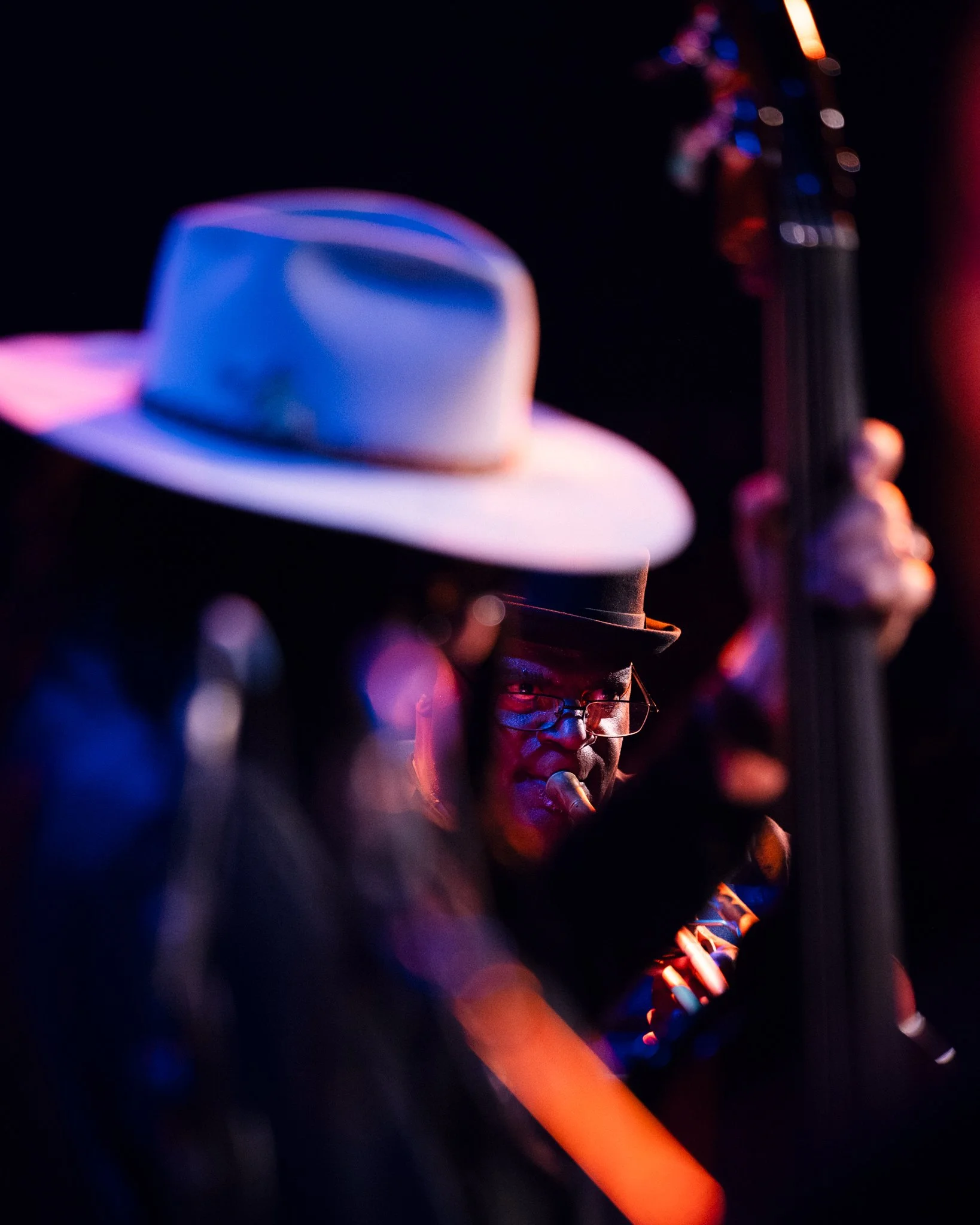 A person playing a guitar in a dark setting, wearing glasses and a fedora hat, with colorful stage lighting.