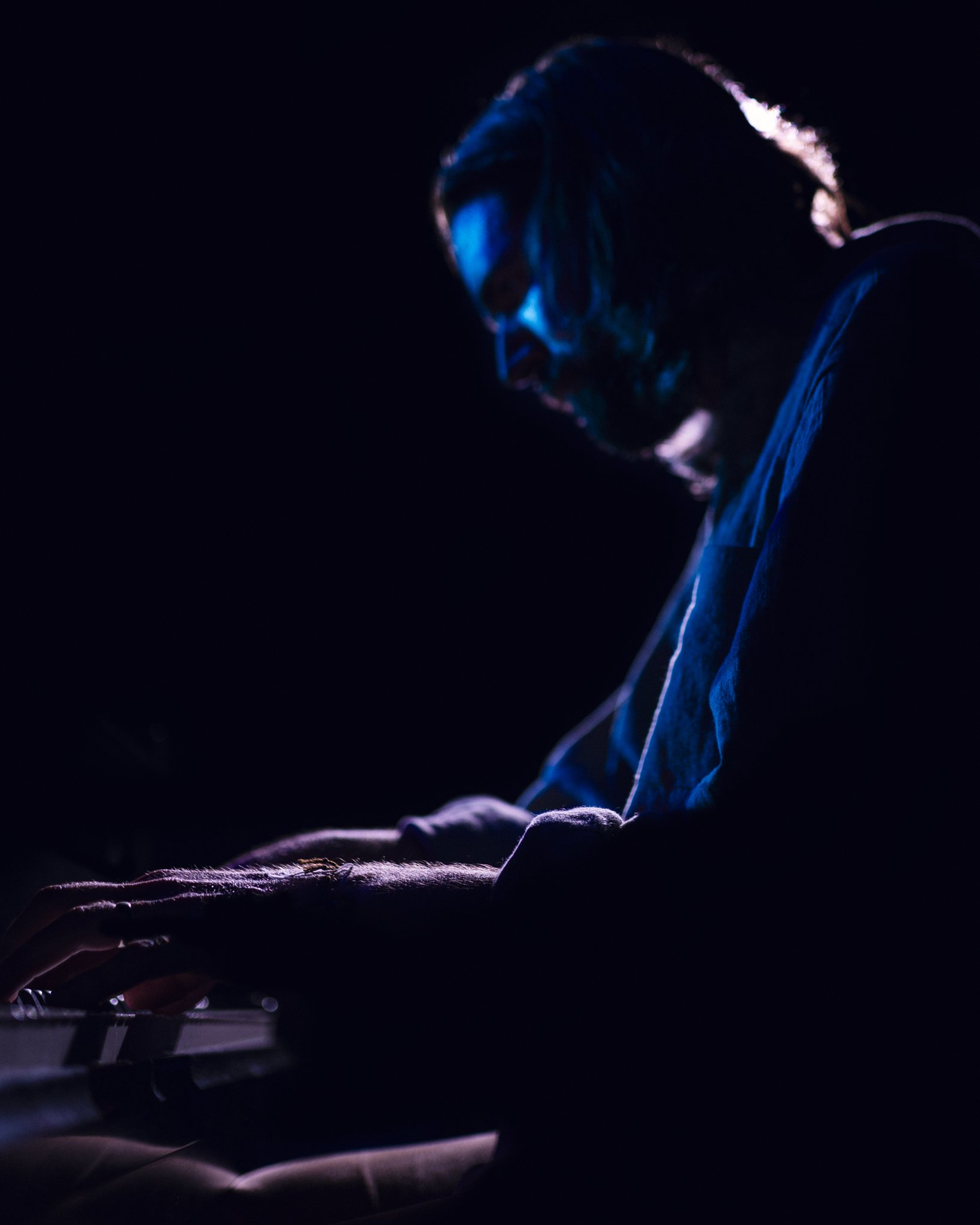 A man with long hair and glasses playing piano in a dark setting, illuminated by a soft blue light.