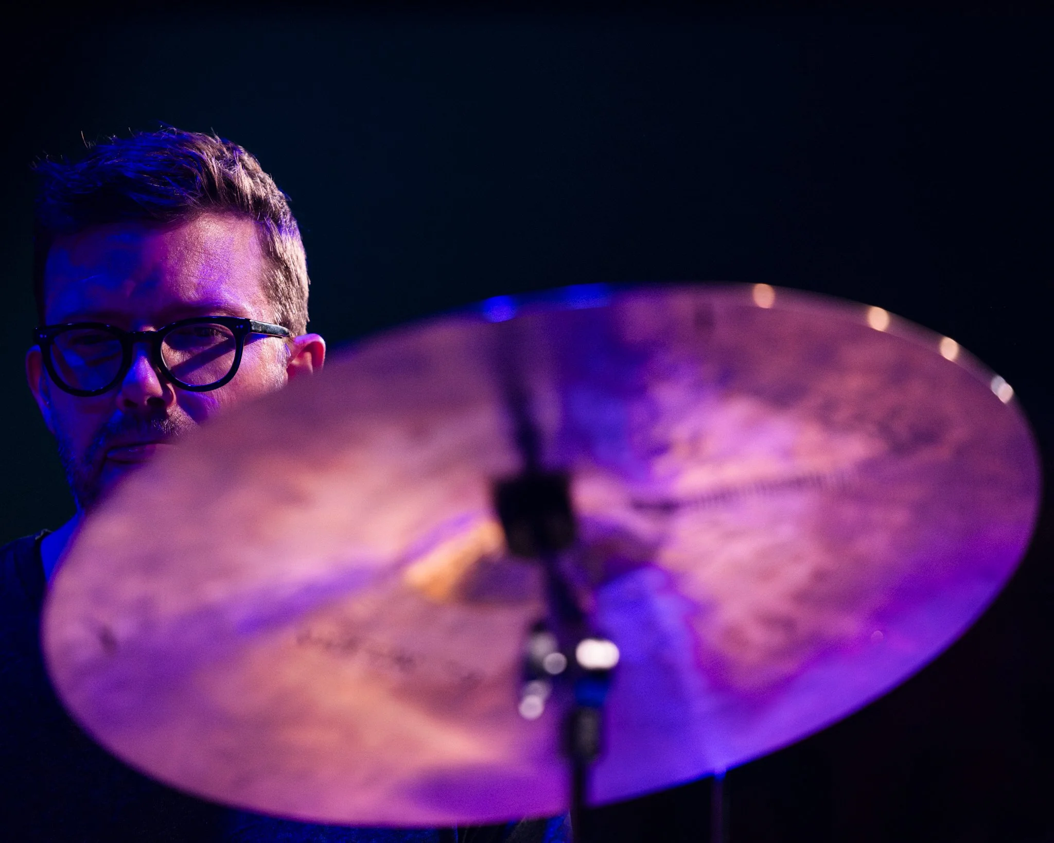 A man with glasses playing a cymbal on a drum set, with purple and blue stage lighting.