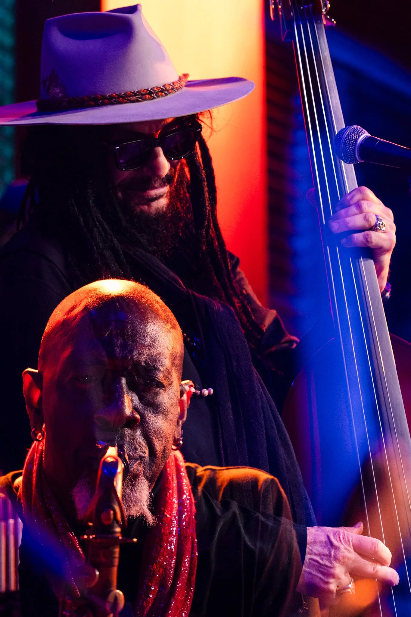 Two musicians performing on stage, one playing the upright bass and the other a saxophone, under colorful stage lights.