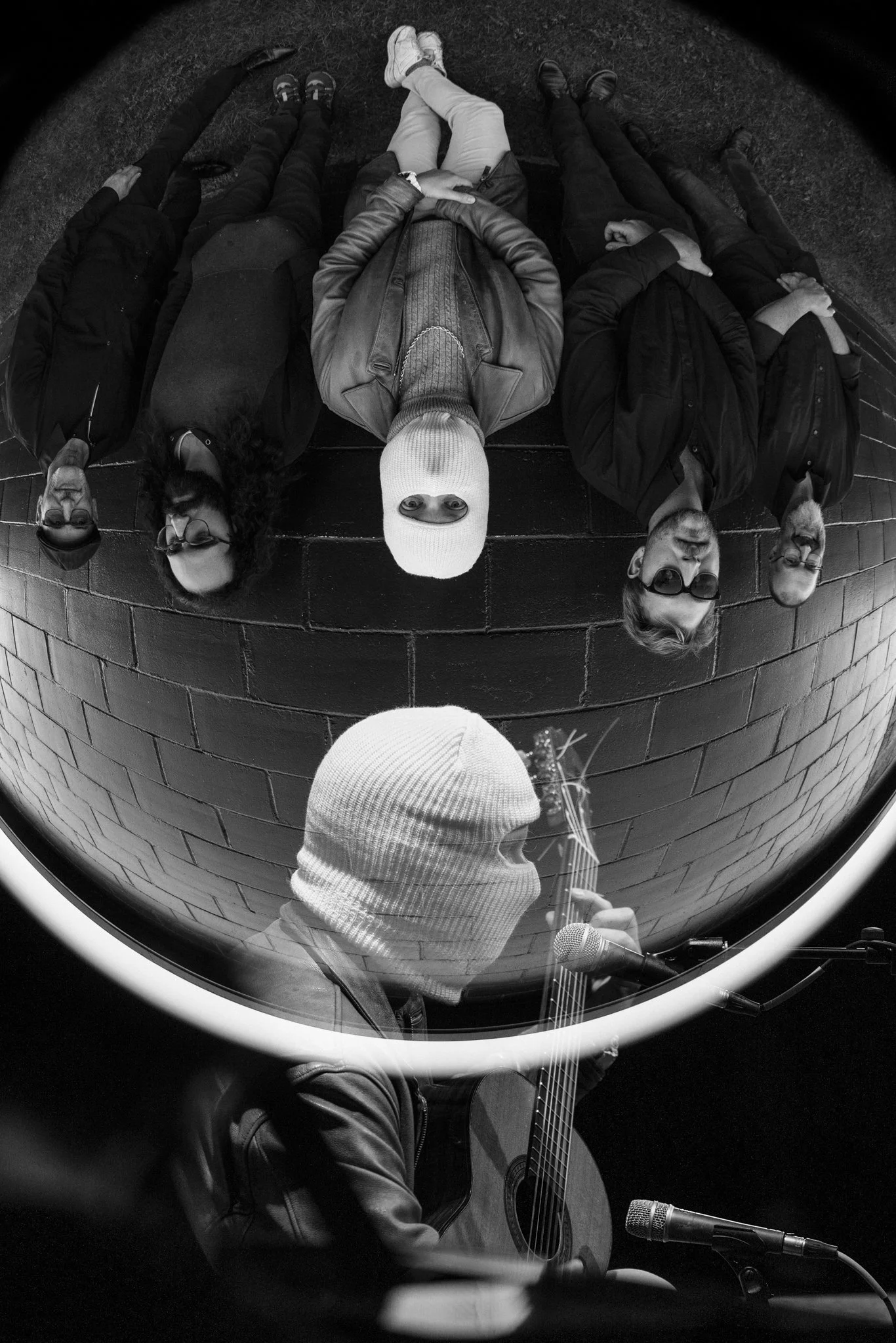 A person in a white knit beanie and leather jacket is playing guitar and singing into a microphone, with a circular mirror reflecting five people sitting against a black brick wall. The reflected group includes three women and two men, all wearing da