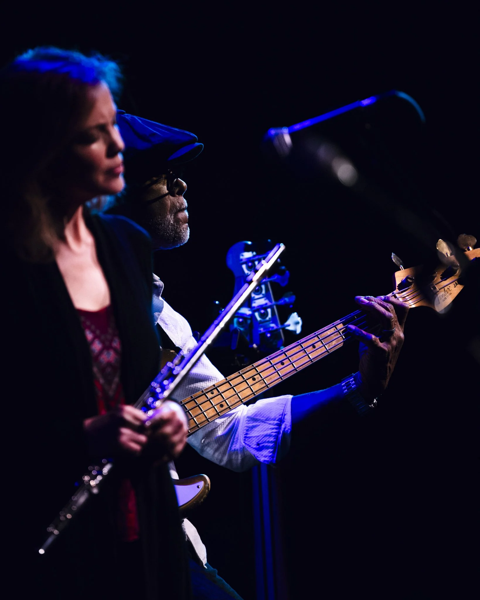 Two musicians on stage, one playing a violin and the other playing a bass guitar, under blue stage lighting.