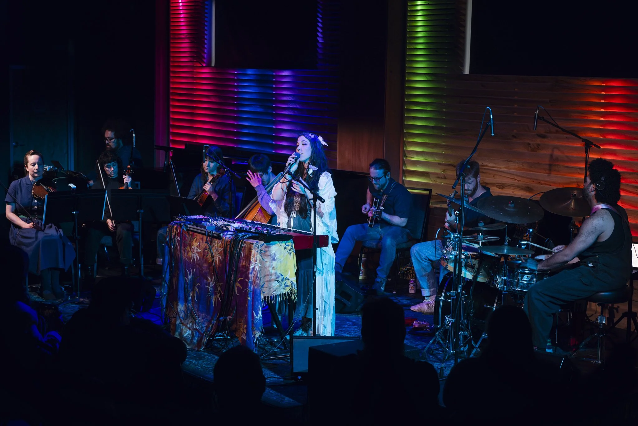 Musicians performing on stage with colorful lighting, including a female singer at the center, surrounded by guitarists, violinists, and drummers.