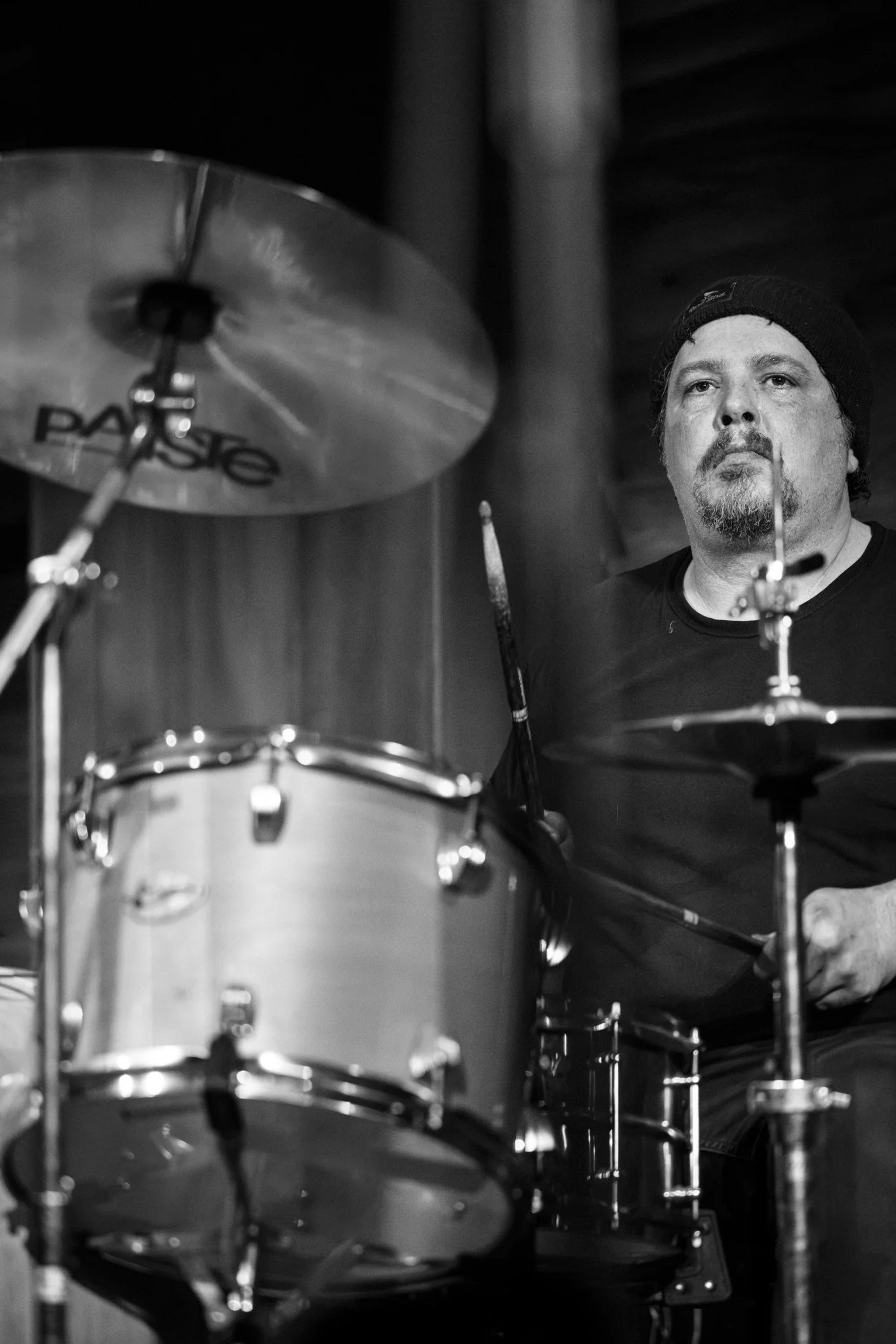A black and white photo of a man wearing a beanie playing drums with a focused expression.