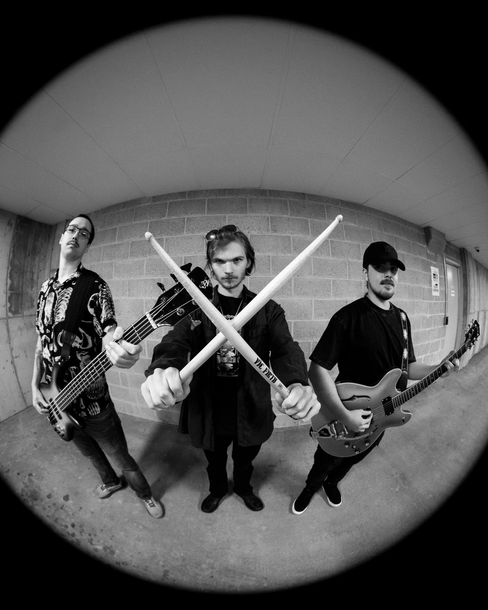 Three young men with musical instruments posing against a brick wall, with one holding drumsticks crossed in front of him, all looking at the camera in black and white.
