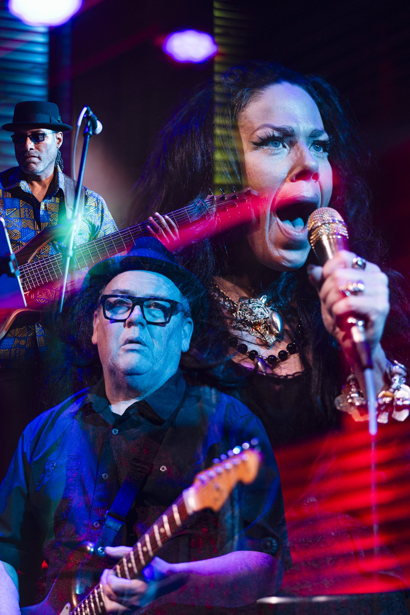 A lively musical performance with a female singer passionately singing into a microphone, accompanied by two male guitarists, one with glasses and a hat, and the other wearing a black hat, all illuminated by colorful stage lighting in a vibrant setti