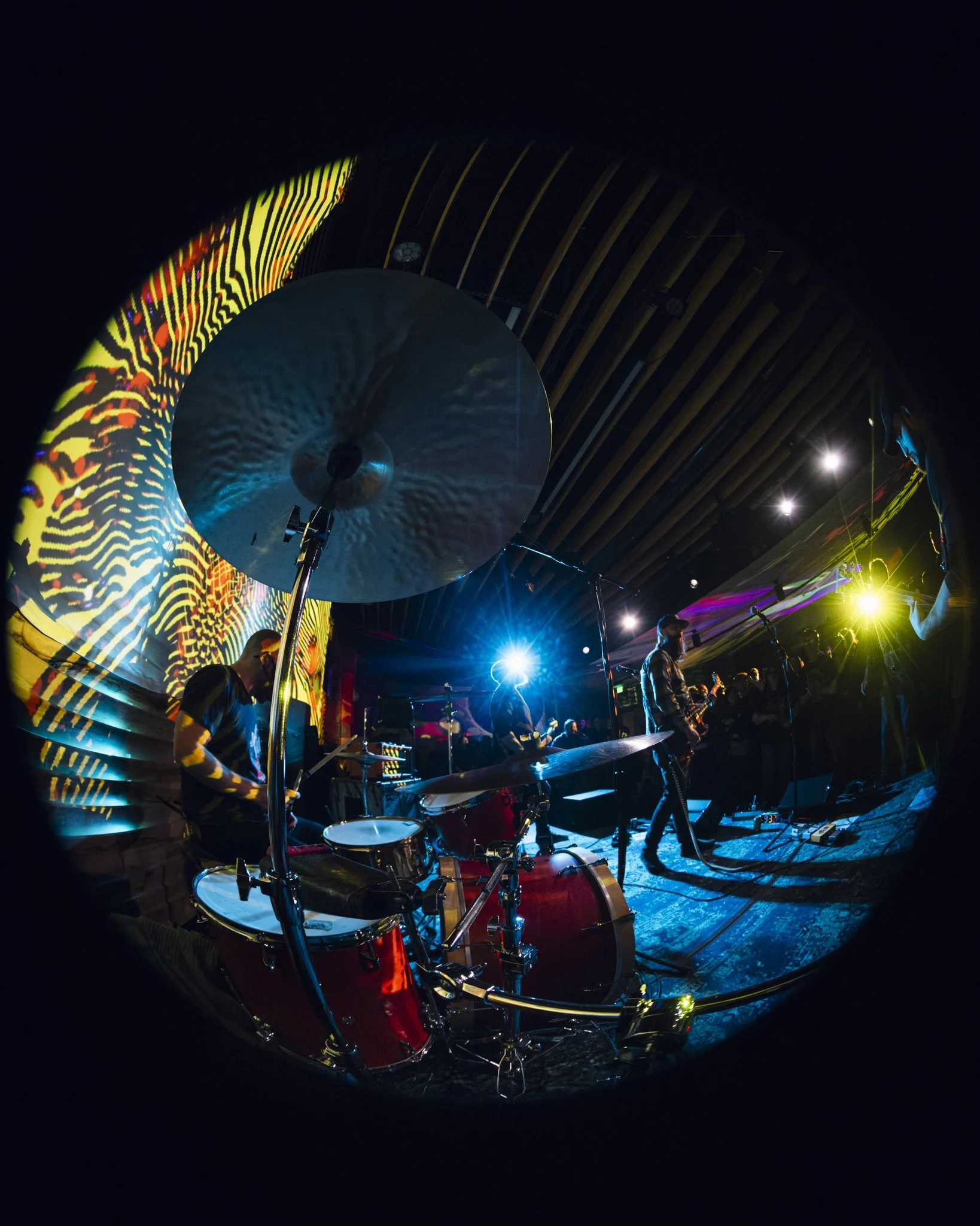 A live band performing on stage with colorful lighting and visual projections, including a drummer, guitarists, and a microphone.