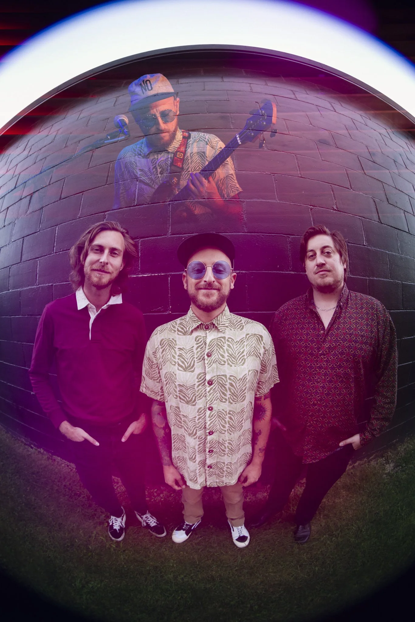 A band poses in front of a brick wall, with a performer playing guitar seen through a circular glass or lens above them.
