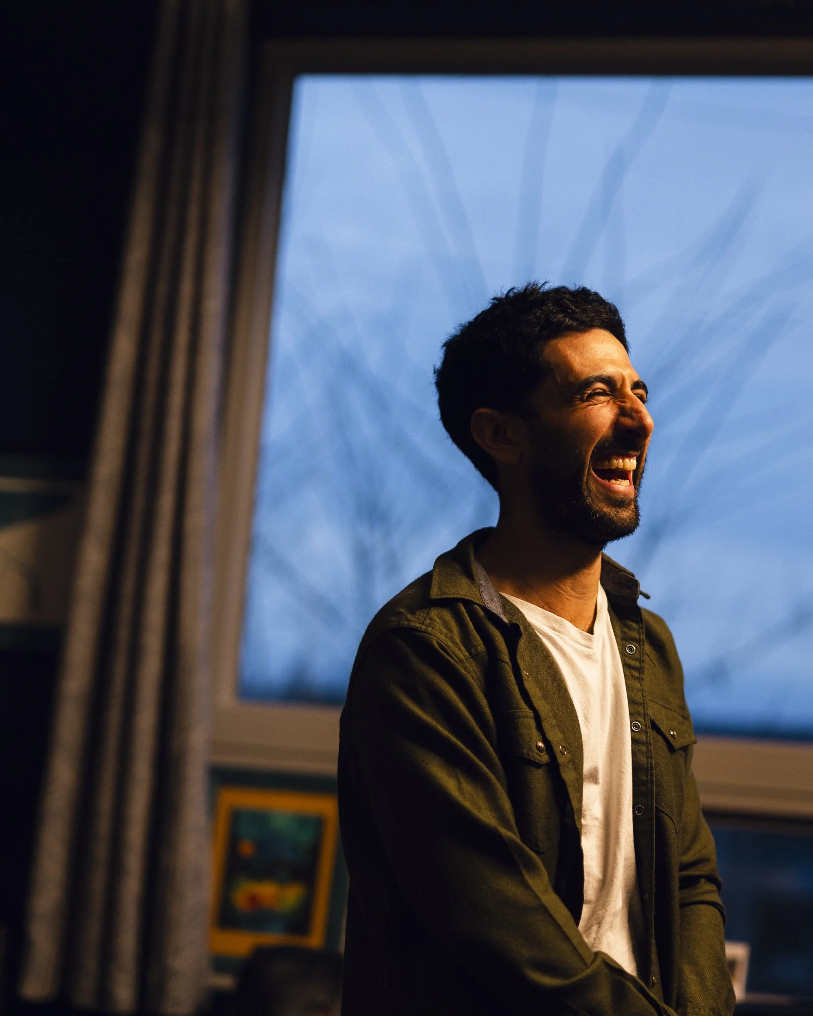 A man standing indoors near a window, laughing or smiling with his eyes closed, at dusk or nighttime outside.