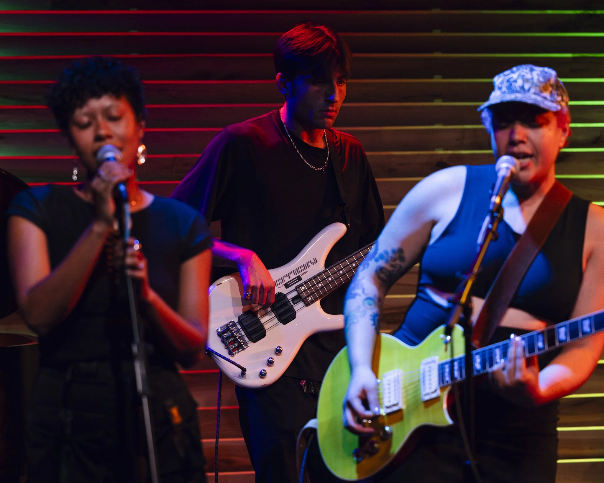 Three musicians performing on stage with a wooden background. The woman on the left is singing into a microphone, the man in the center is playing a white bass guitar, and the woman on the right is playing a yellow-green electric guitar and singing i