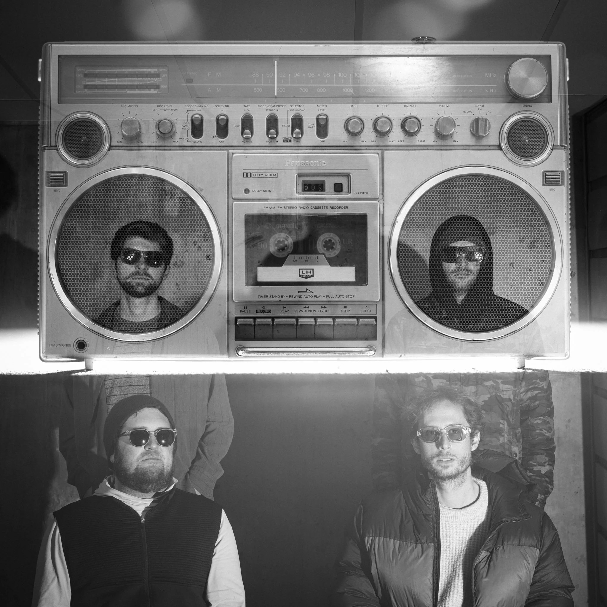 A vintage boombox with two men in hoodies and sunglasses standing in front of it, one on each side, with the boombox positioned above them.