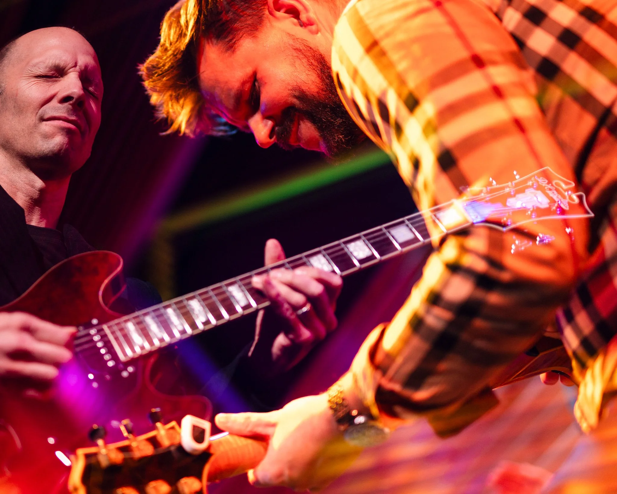 Two men playing electric guitar on stage, with one wearing a black shirt and the other wearing a plaid shirt, both focused on their performance in a vibrant, colorful setting.