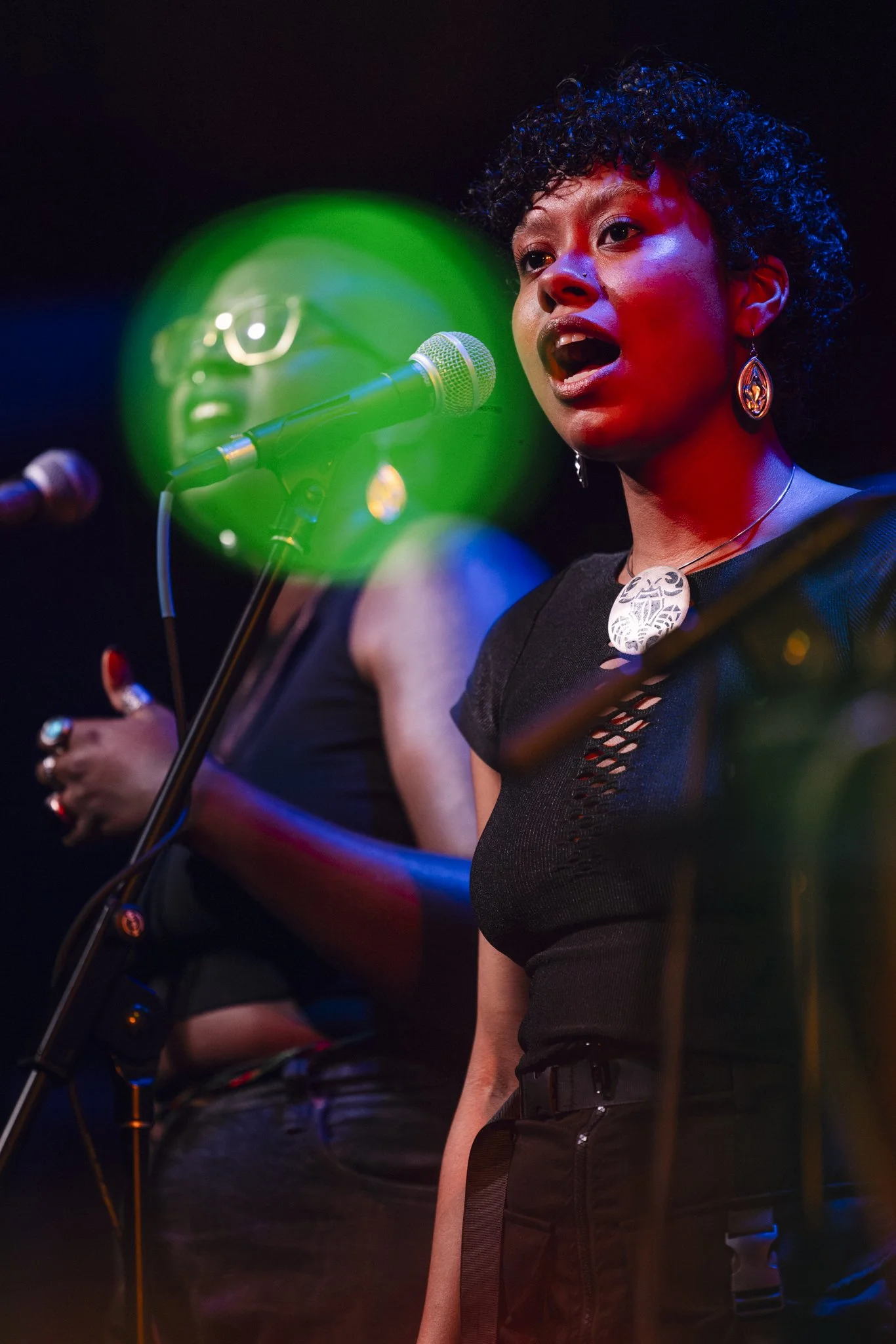 Two women singing into microphones during a performance, with colorful stage lighting.