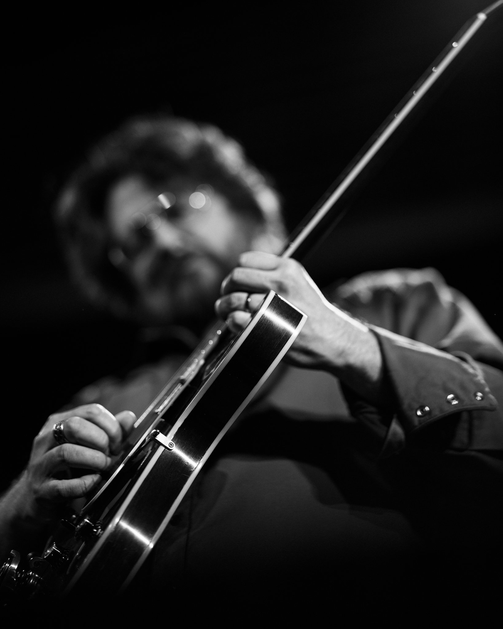 A man playing an acoustic guitar, viewed from a low angle, in black and white with focus on the guitar and blurred face.