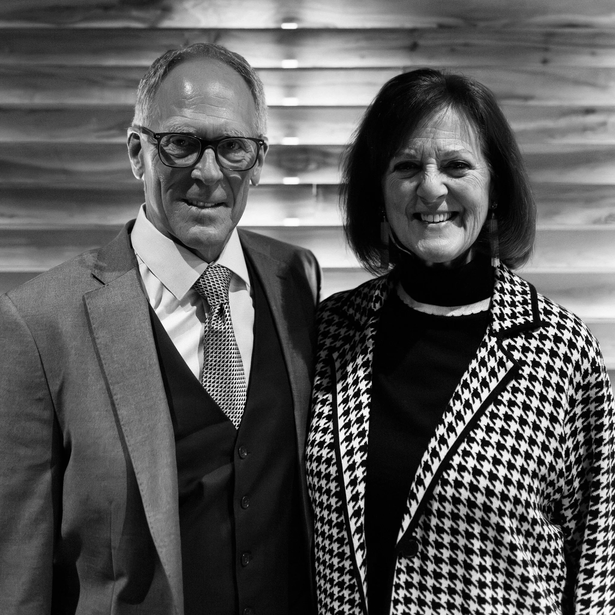 A black and white photo of a man in a suit and a woman in a patterned jacket standing together and smiling.
