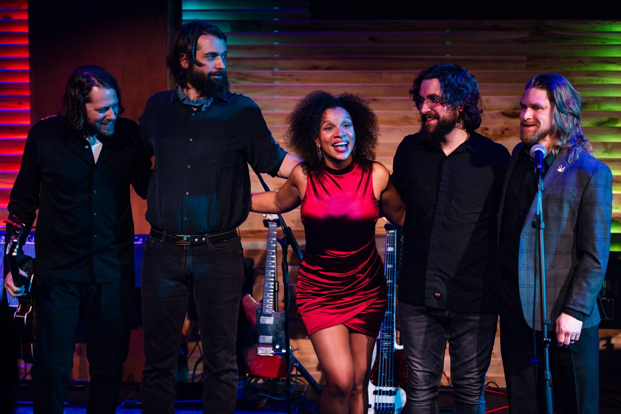 A group of five musicians on stage, smiling and holding each other, with musical instruments in the background, in a colorful lit venue.