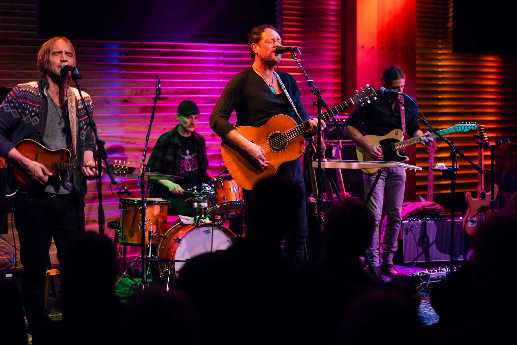 A band performing live on stage with four members. Two musicians are playing acoustic guitars and singing into microphones, one is on the drums, and the other on an electric guitar. The stage has colorful lighting with pink and orange hues.