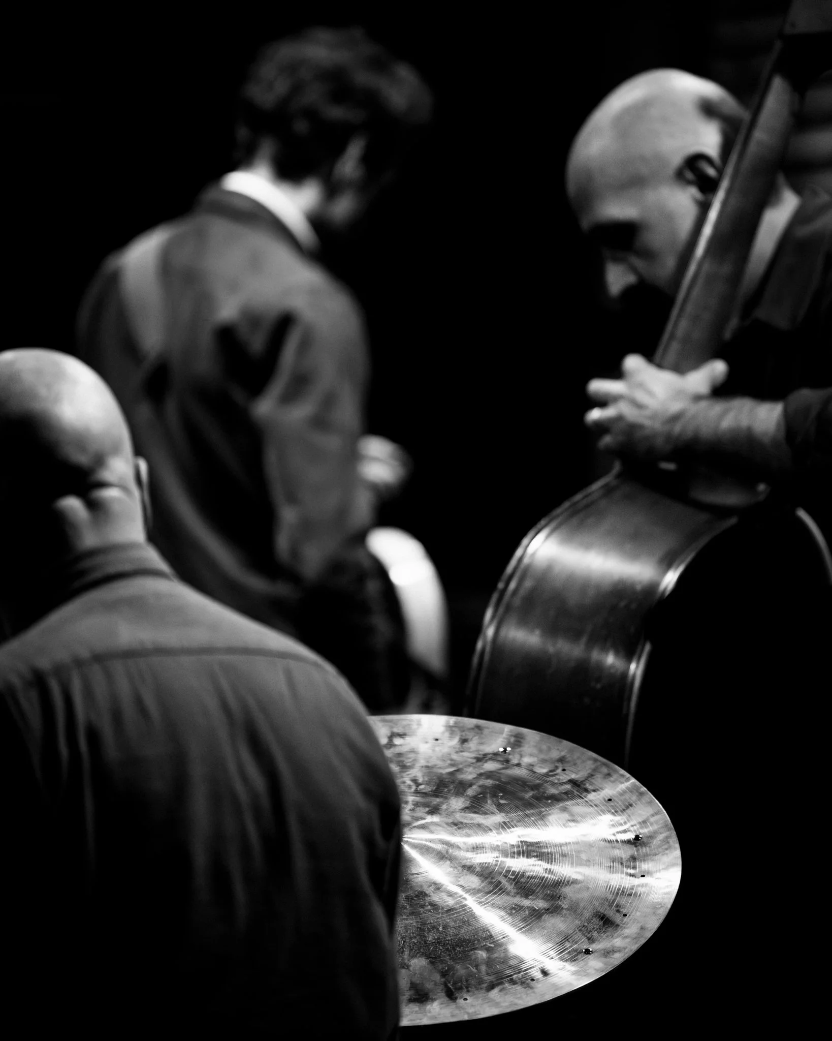 Black and white photo of three musicians, one playing drums, another playing double bass, and the third appears to be conducting or resting, in a dark setting.
