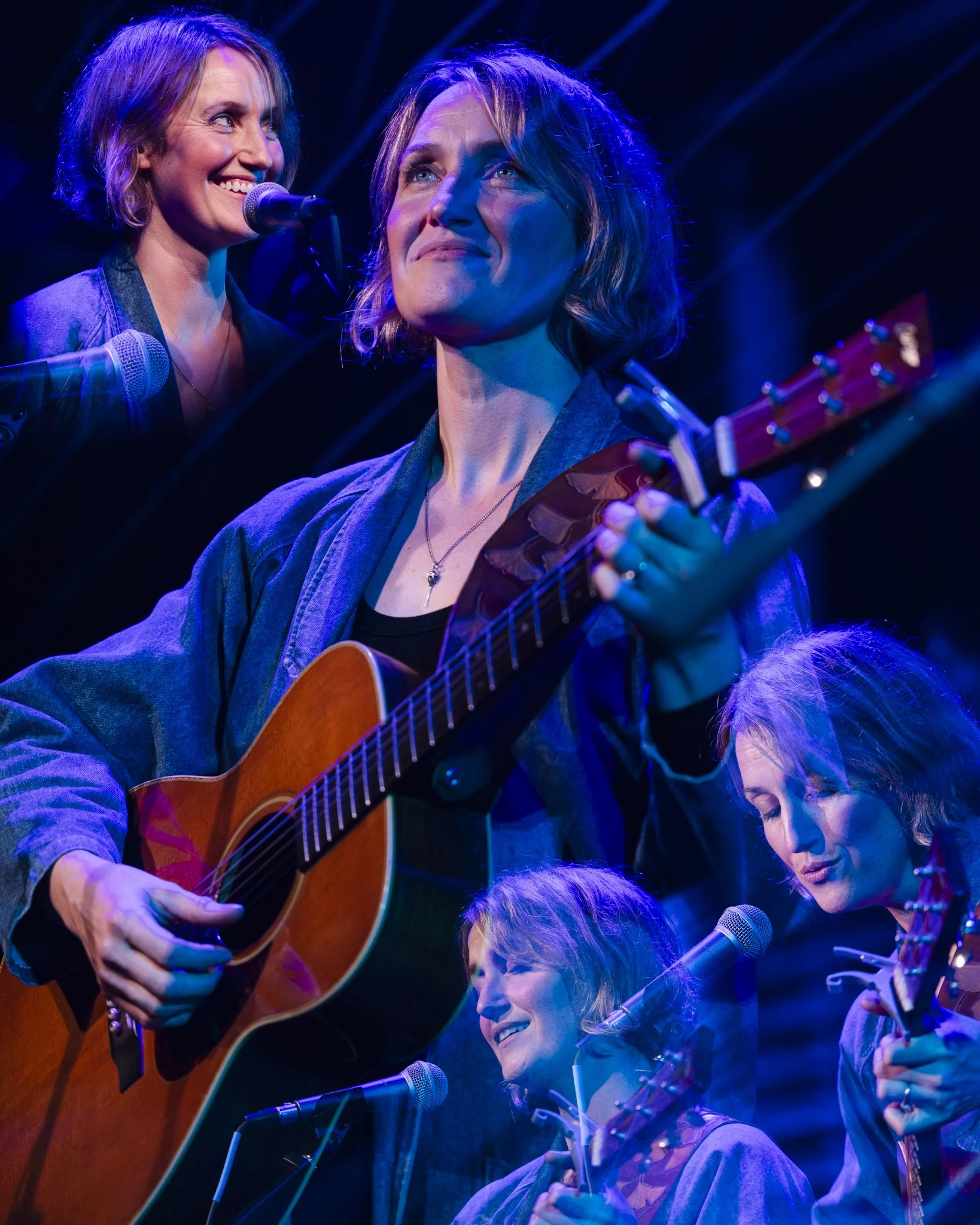 Multiple women performing music on stage, one playing guitar and singing, others holding guitars and singing, with stage lighting.