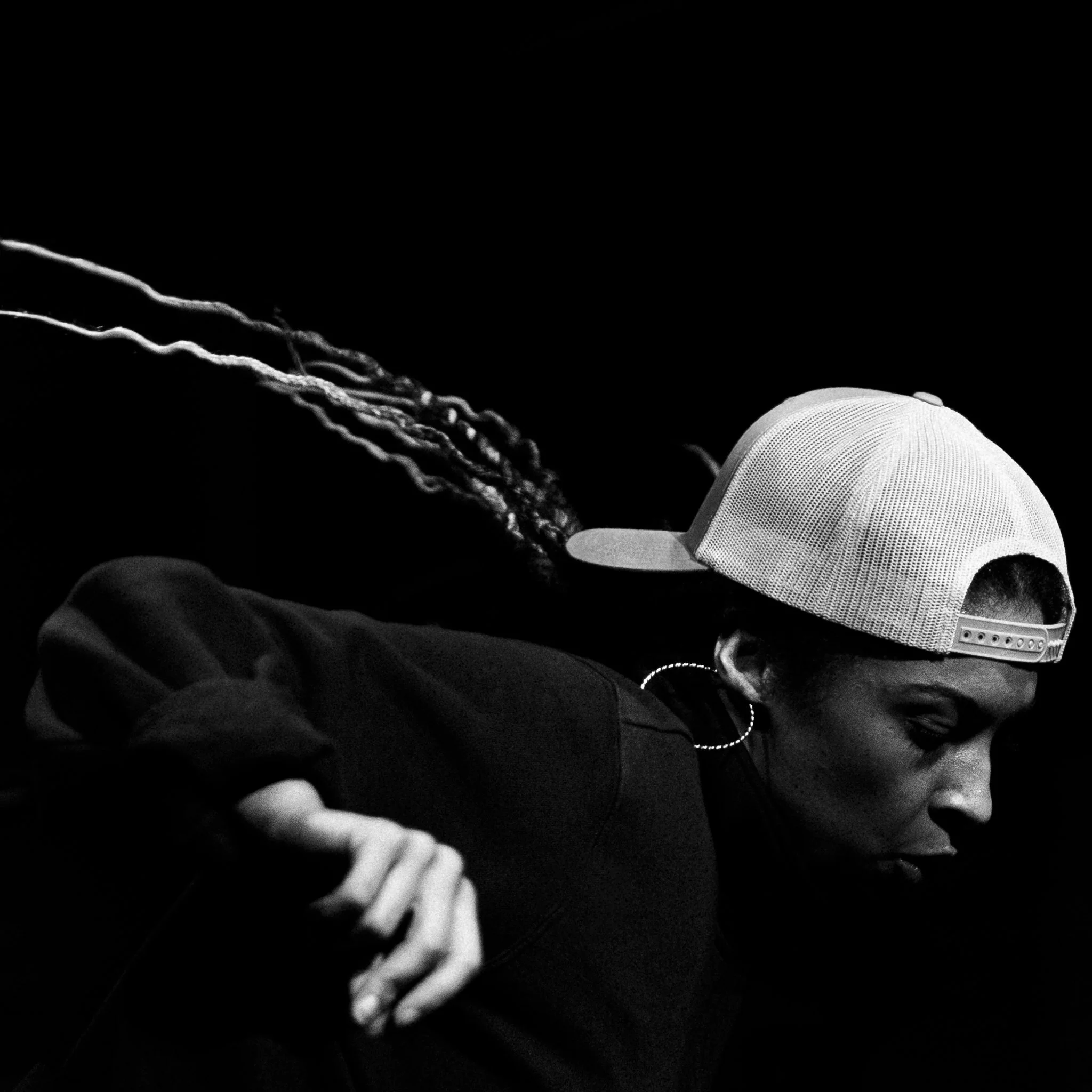 Black and white photo of a person with a backward baseball cap, wearing a hoop earring, and a tattoo sleeve on their arm, looking downward.