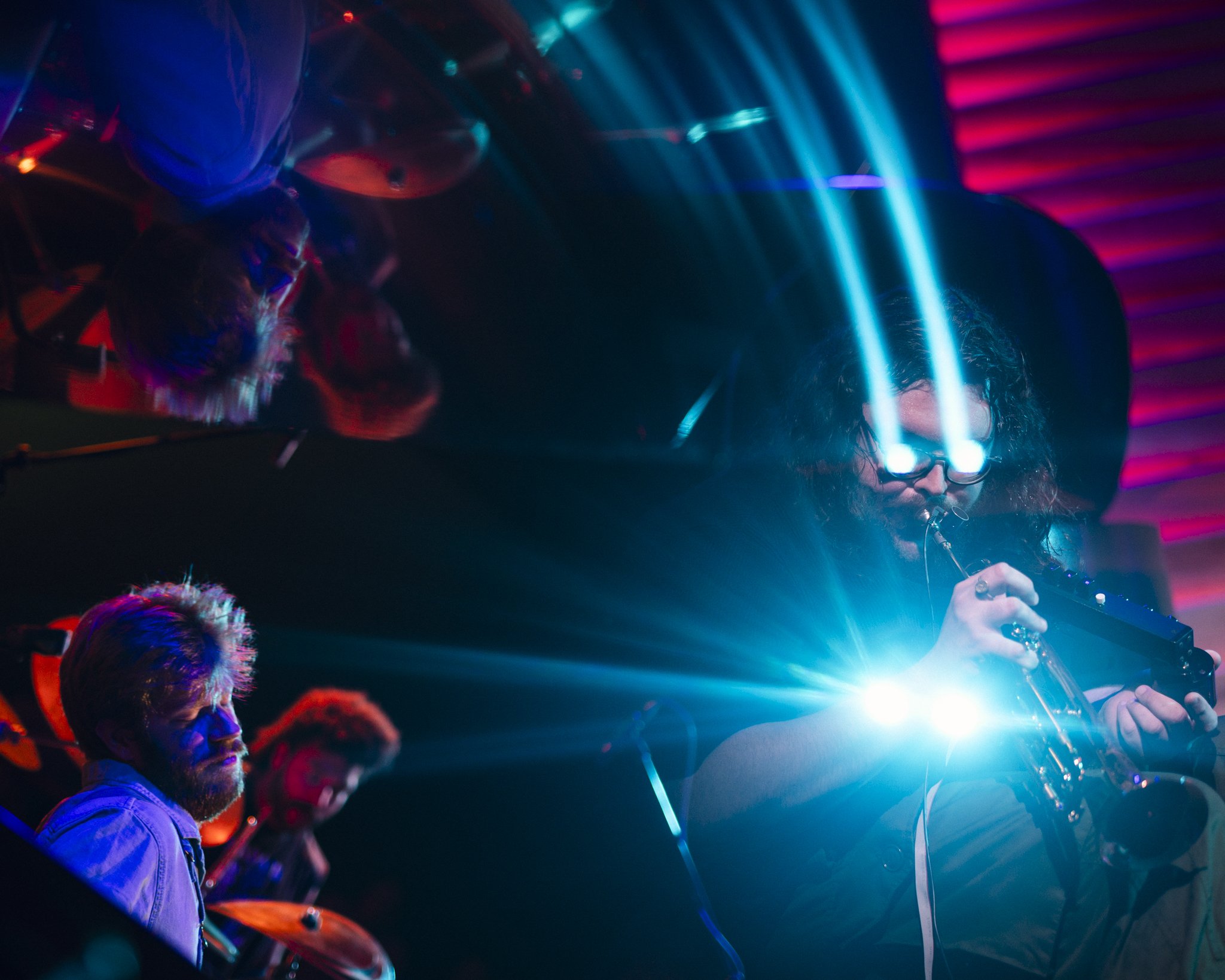 A musician wearing glasses with blue laser beams coming out of the glasses, playing a violin on stage with colorful lights. Two other band members are visible in the background.