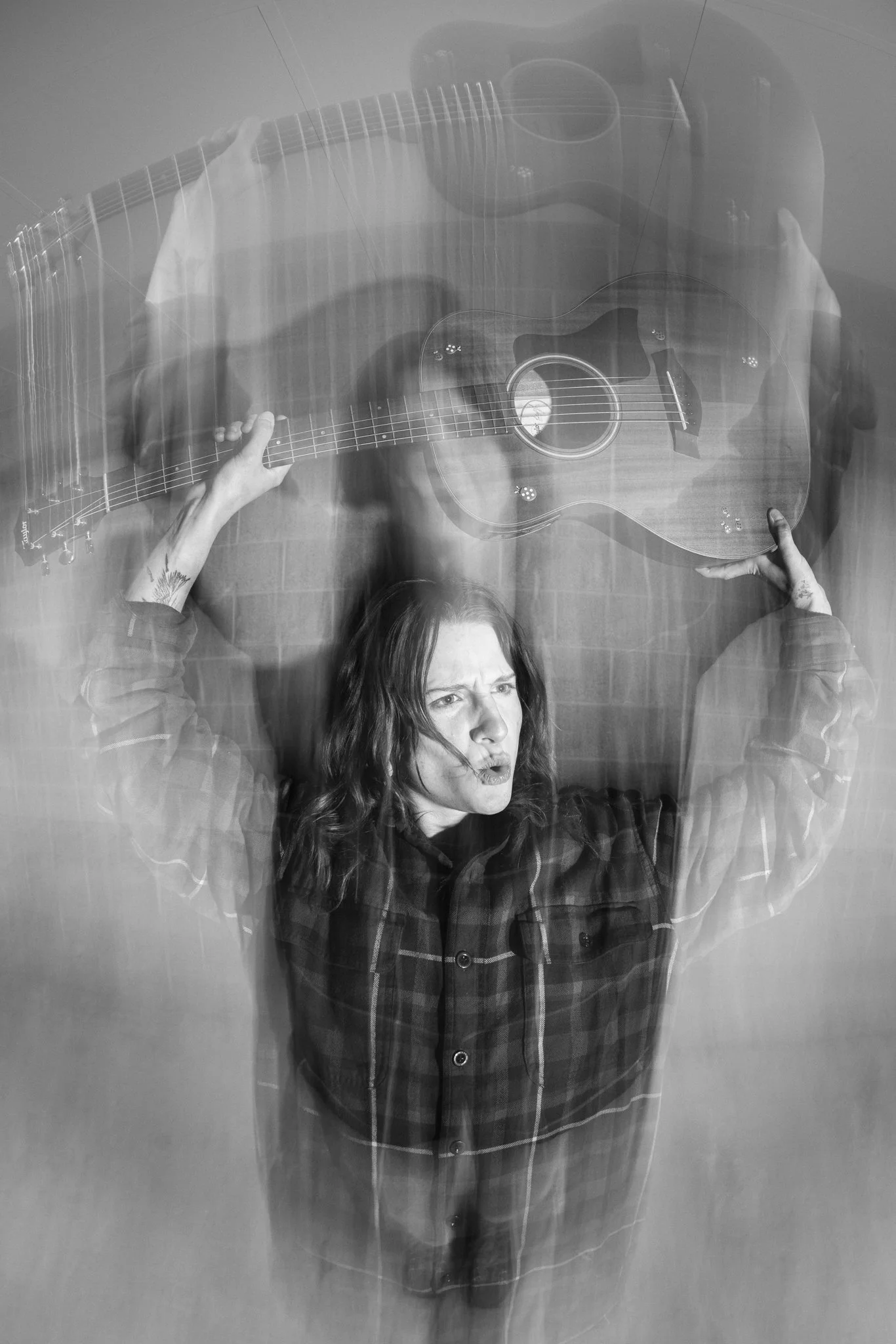 Black and white photo of a woman holding a guitar above her head, with a double exposure effect showing her facial expression and her wearing a checkered shirt.