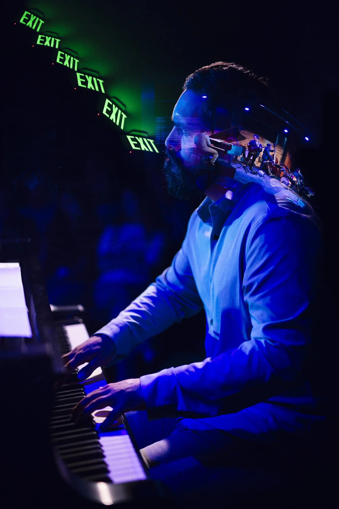 A man playing a piano at a dimly lit jazz club with a reflection of a live band on the glass, and green 'EXIT' signs on the wall.