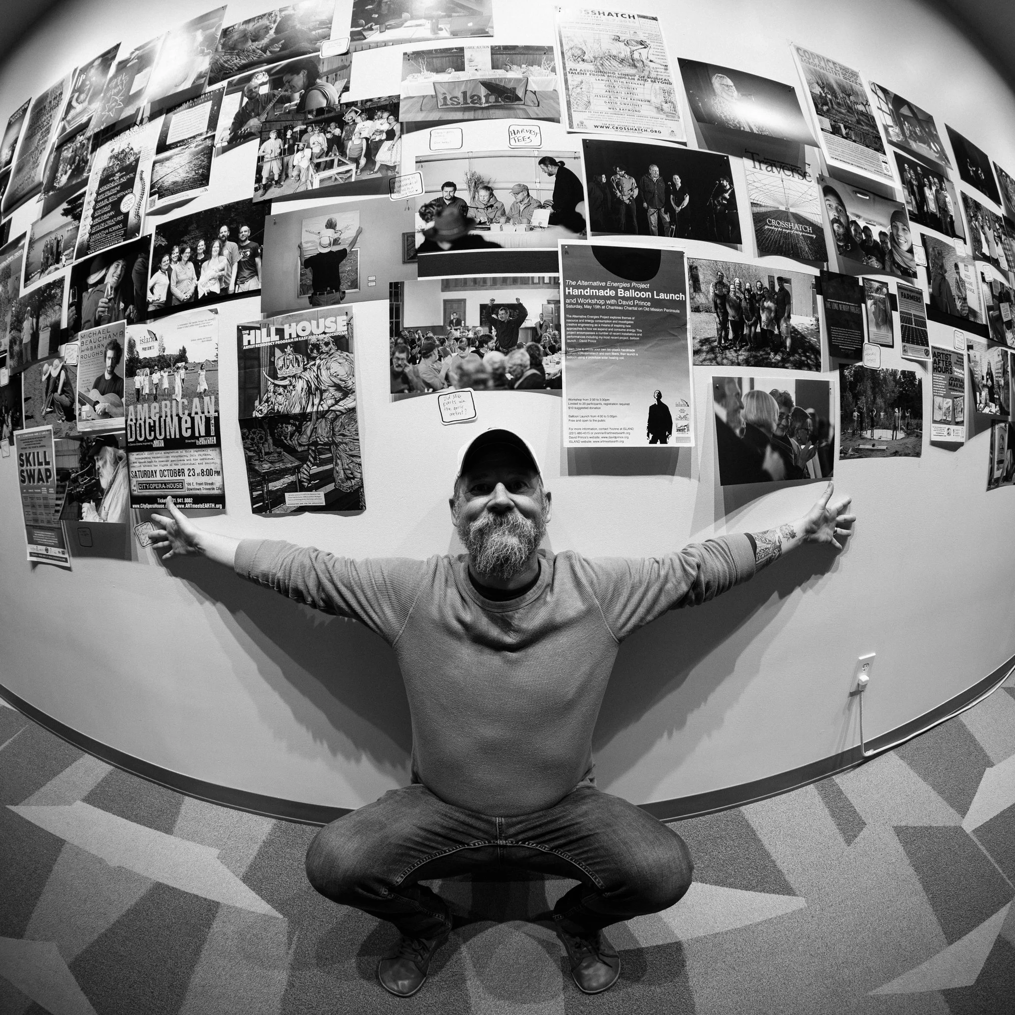 A man with a beard and tattoos poses with arms outstretched in front of a wall covered with photos and posters, in a room with a patterned carpet.