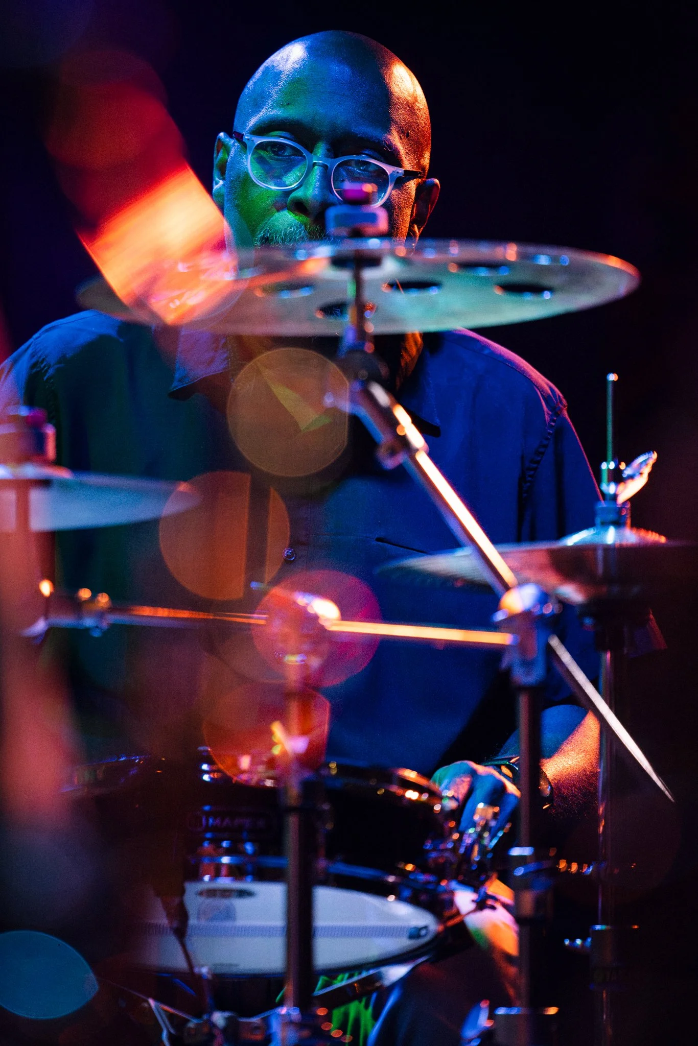 A man wearing glasses playing a drum set, with colorful lighting effects and a dark background.