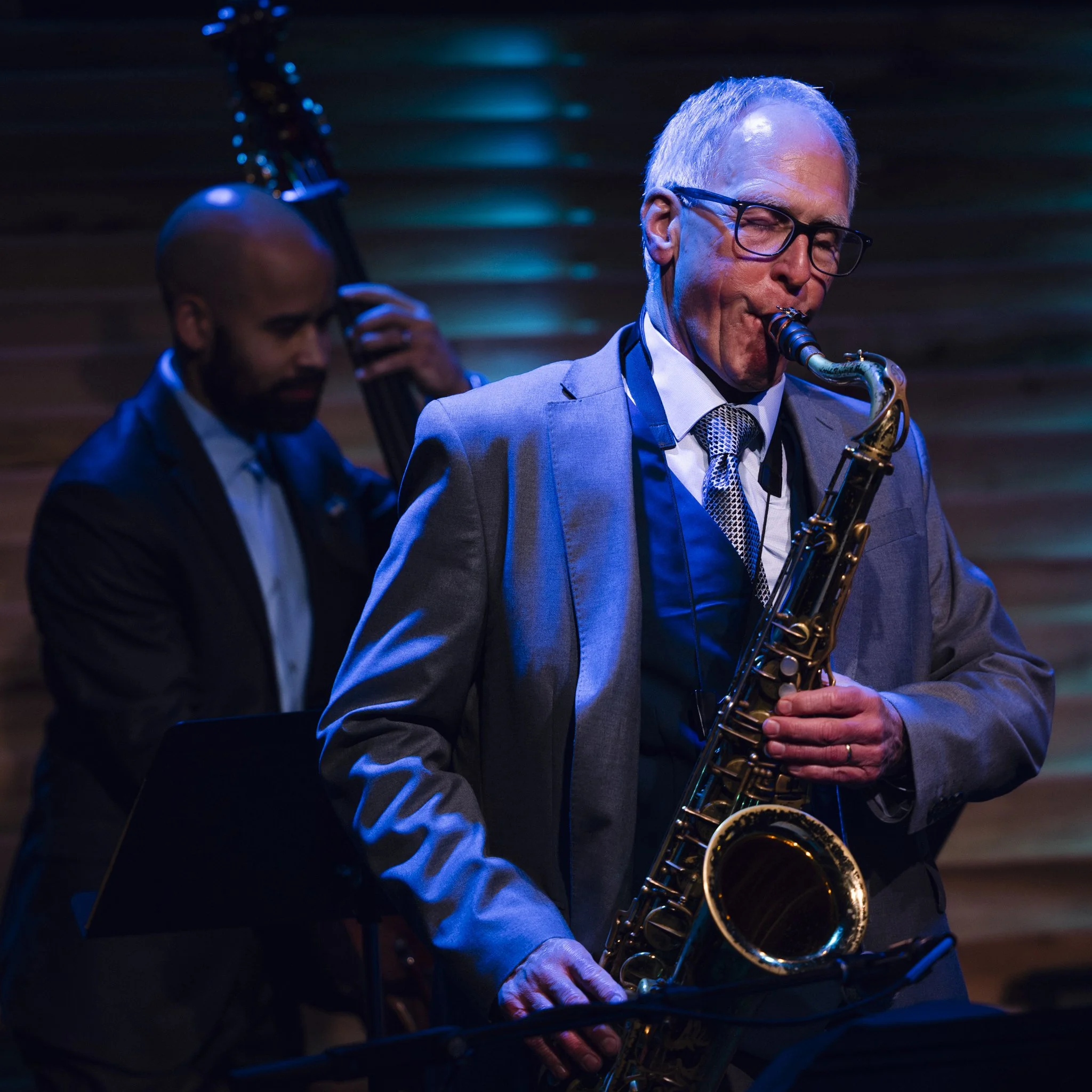 Two jazz musicians performing on stage: one playing a double bass and the other playing a saxophone, with a wooden background and stage lighting.