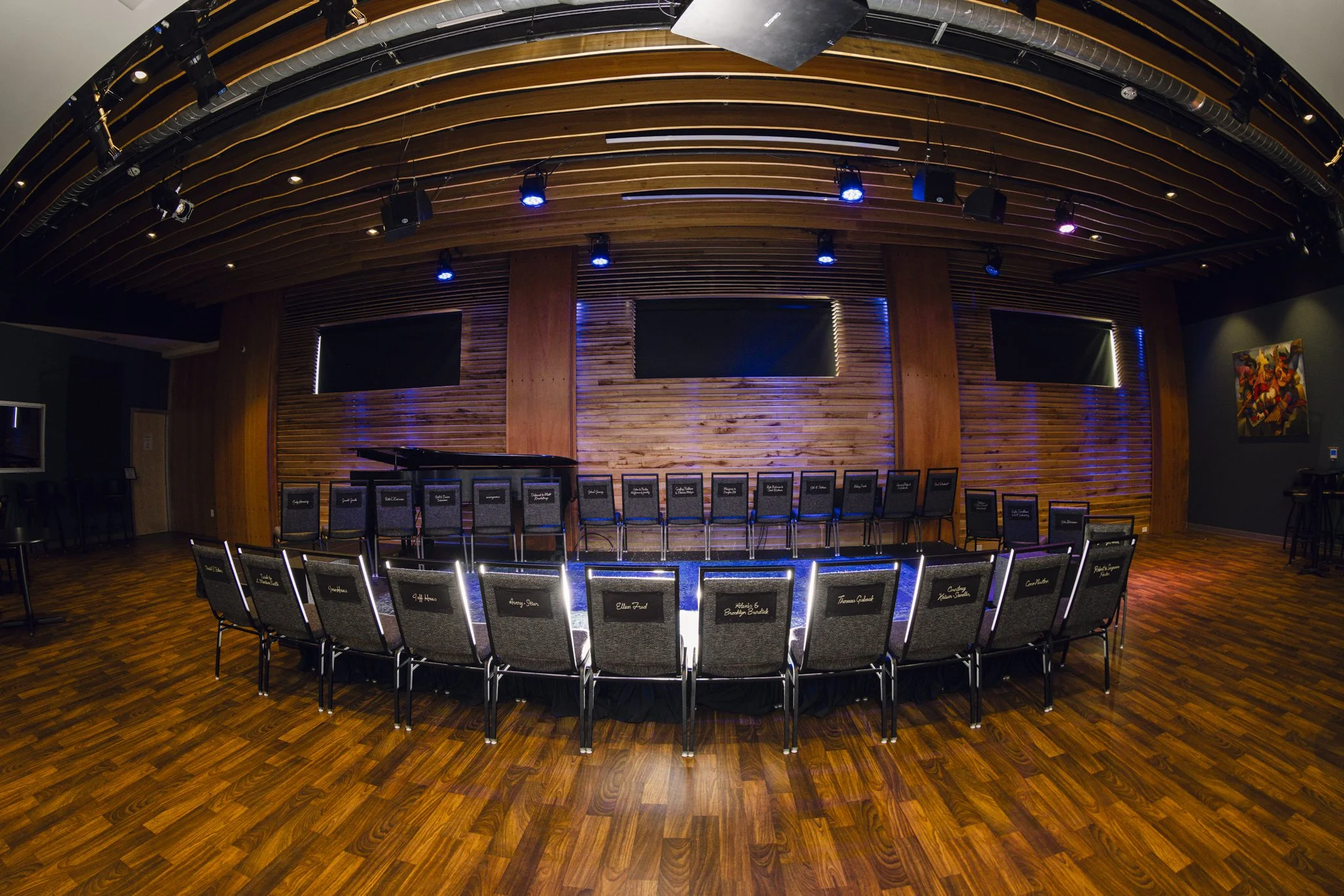 Empty stage with rows of chairs arranged in front, set up for a meeting or event, with a wooden wall background and three large blank screens.