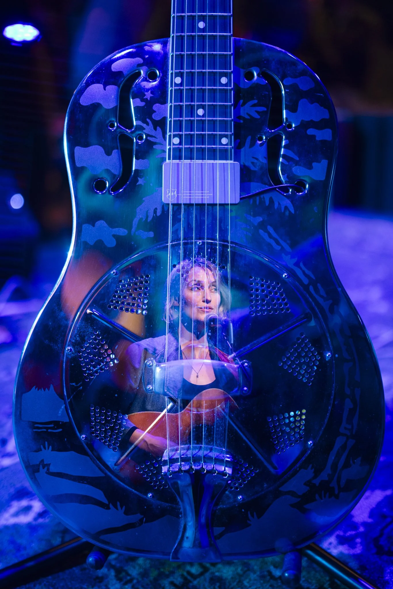 A musician playing an acoustic guitar, seen through a clear plastic guitar-shaped amplifier with a woman reflected on it, in a blue-lit stage setting.