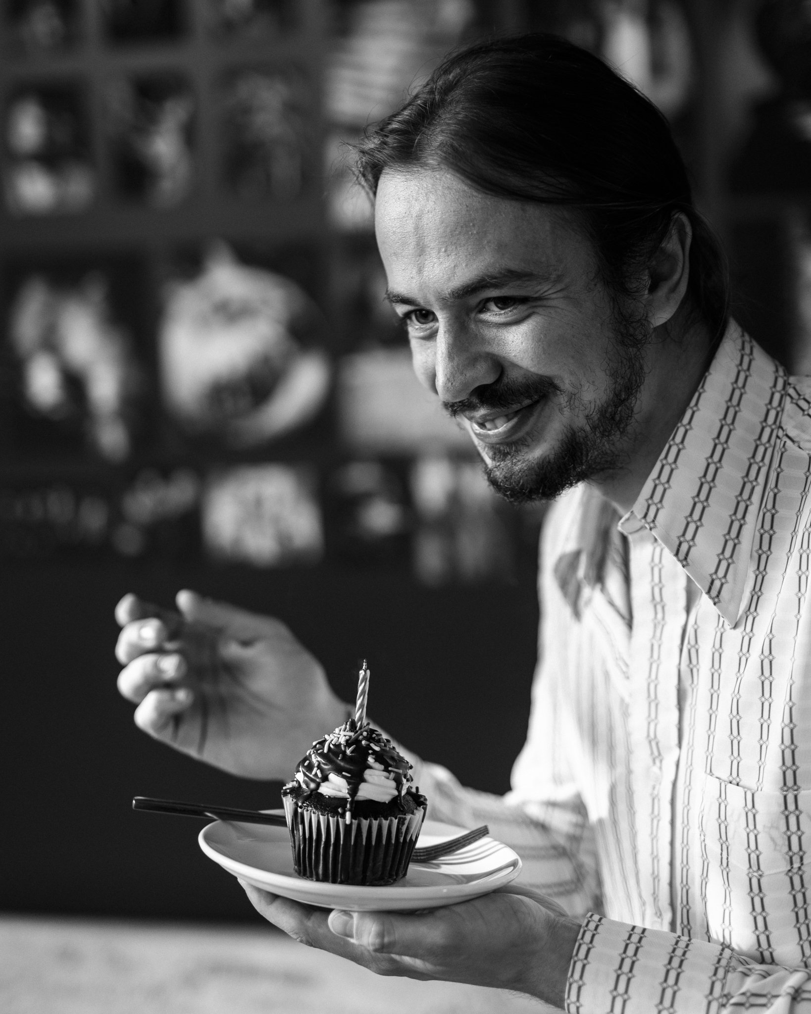 A man with long hair, a beard, and a patterned shirt is smiling and holding a plate with a decorated cupcake that has a lit candle on top.