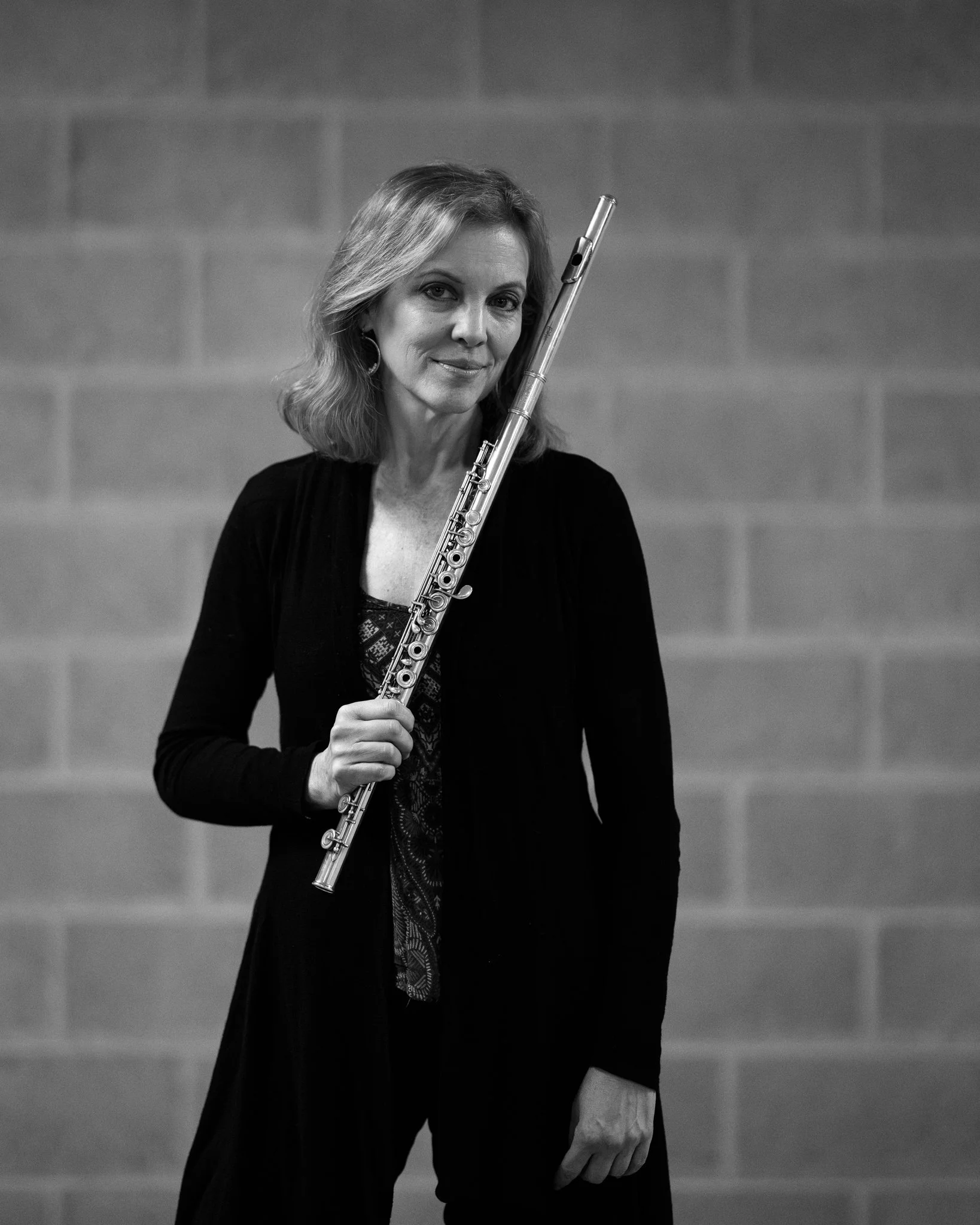 A woman with shoulder-length hair holding a flute, standing against a brick wall.