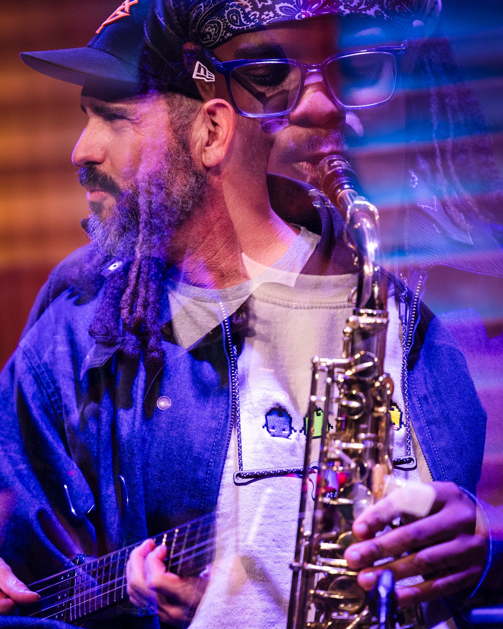 Two musicians, one playing guitar and the other playing saxophone, performing together on stage with colorful lighting.
