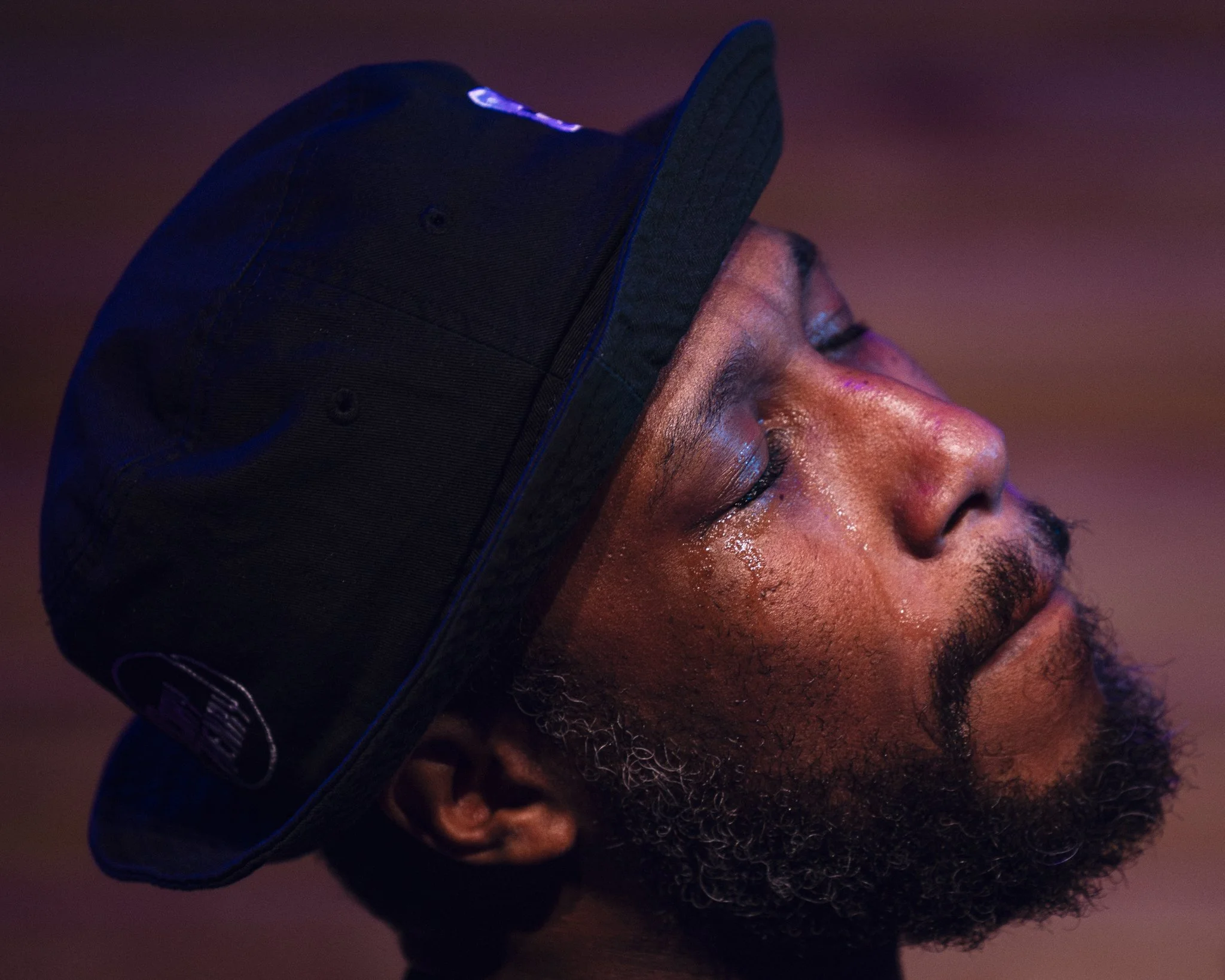 A man with closed eyes, a beard, and tears on his face, wearing a black cap, against a purple and pink blurred background.