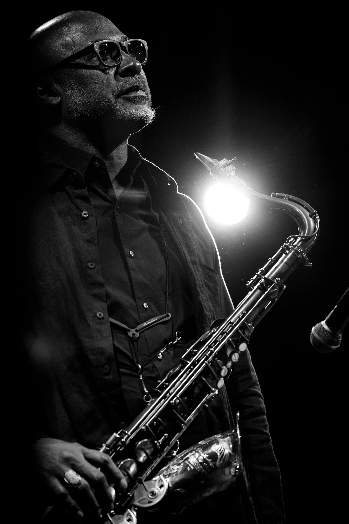 A black and white photo of a man with glasses playing a saxophone on stage with a bright light behind him.