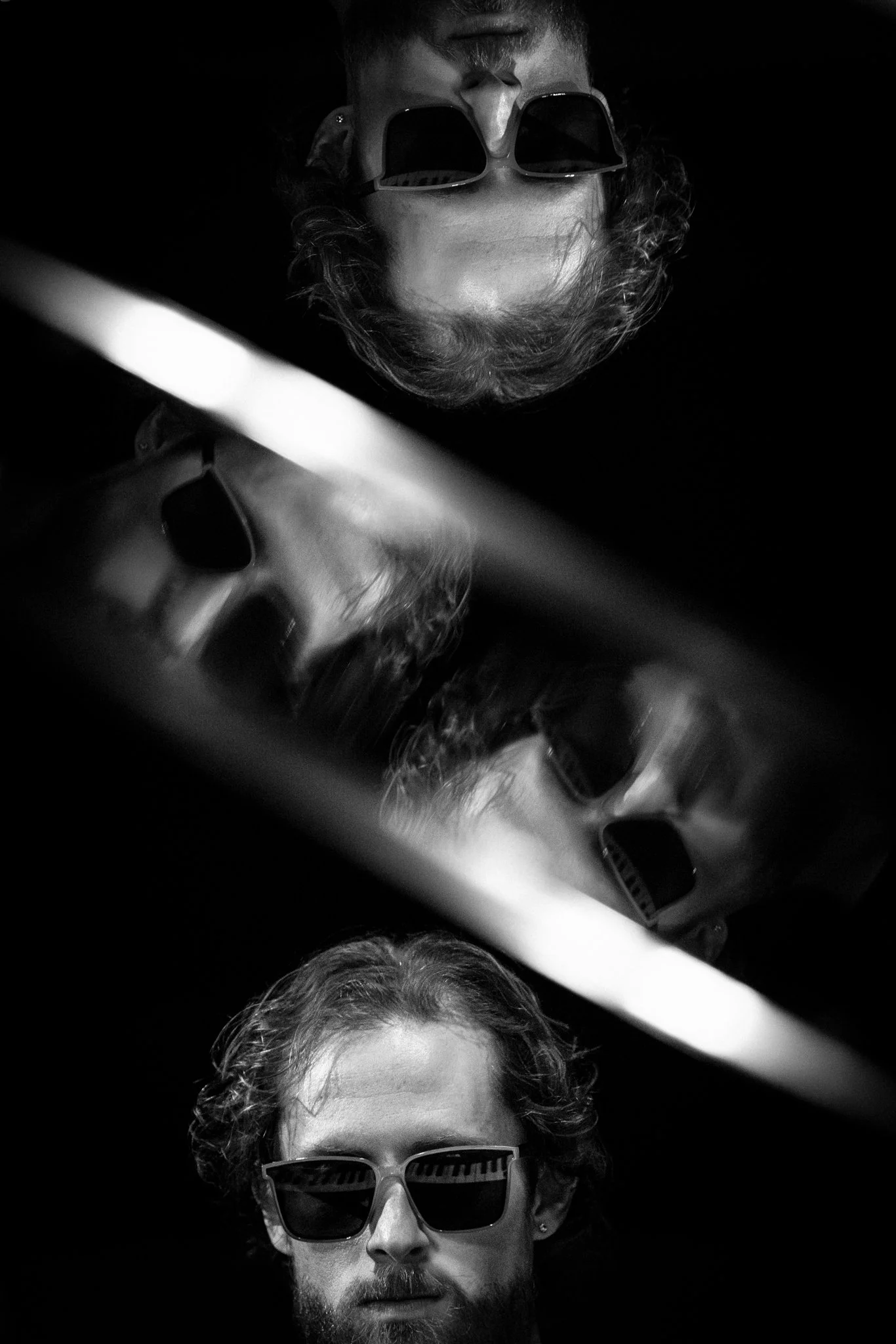 A black and white portrait of a man with curly hair and a beard, wearing sunglasses, with his reflection visible on a curved, shiny surface above.