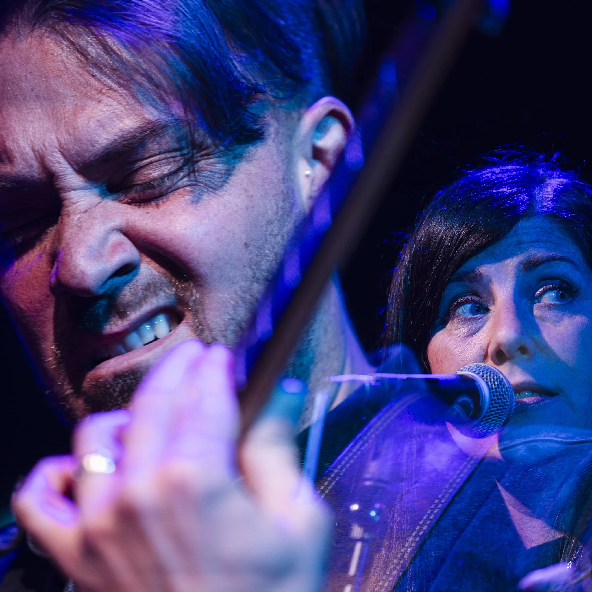 Close-up of a male musician playing a violin with a facial expression of intense emotion and a female singer singing into a microphone, both under blue and purple stage lighting.