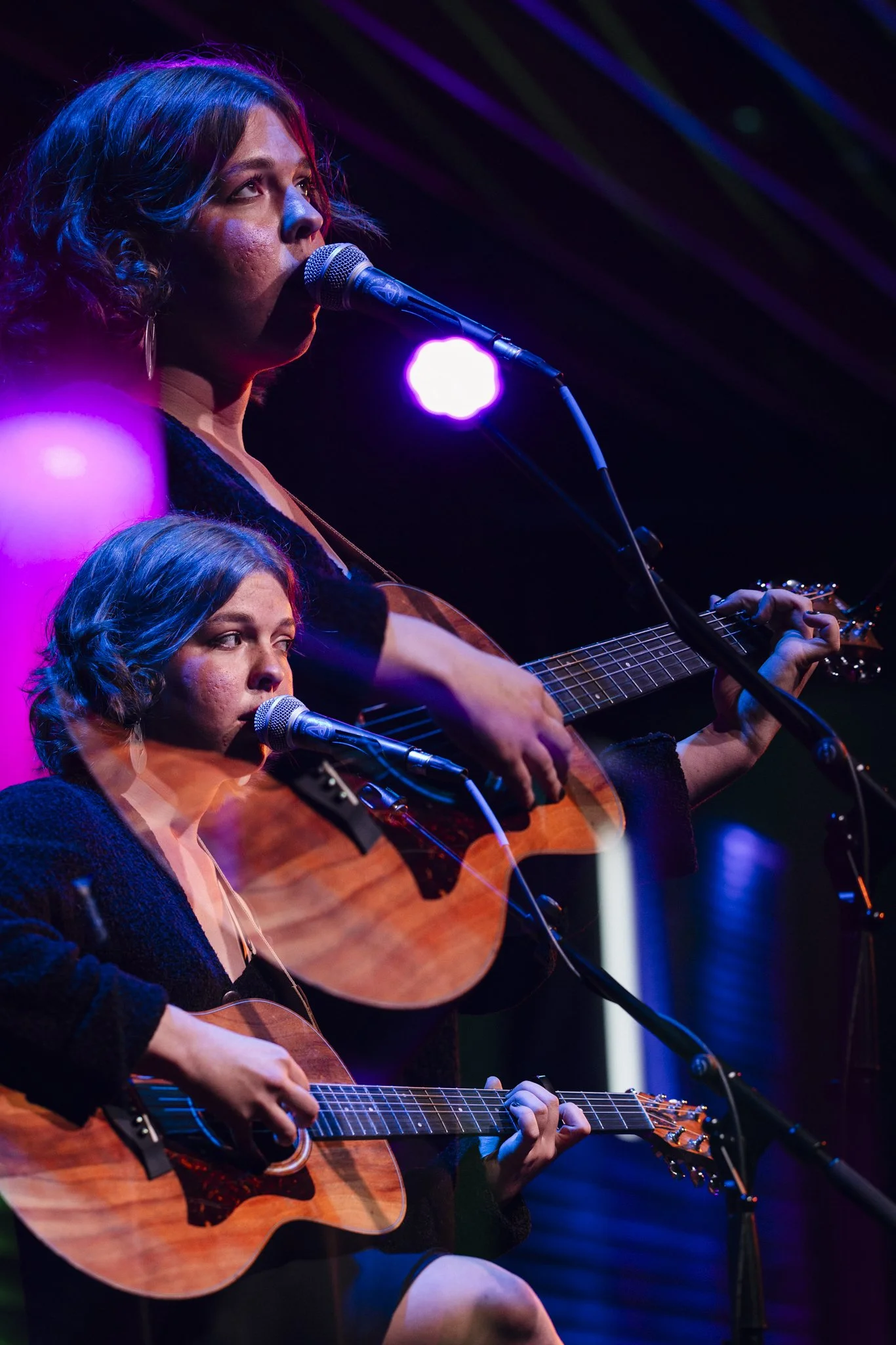 Two women performing on stage with acoustic guitars, singing into microphones, under colorful stage lighting.