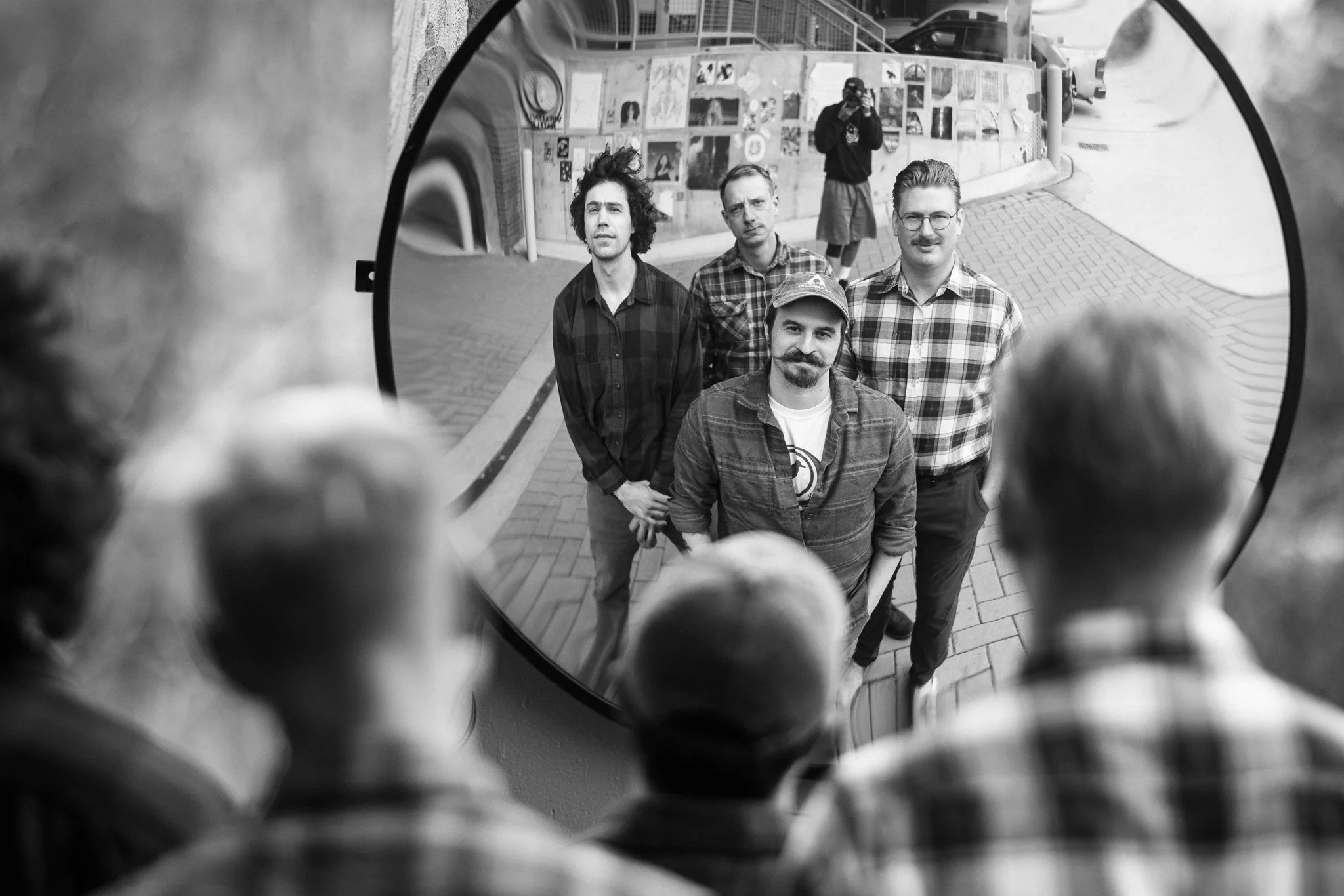 A black-and-white photo of four men standing on a sidewalk, viewed through a round mirror, with two people in the foreground observing them.