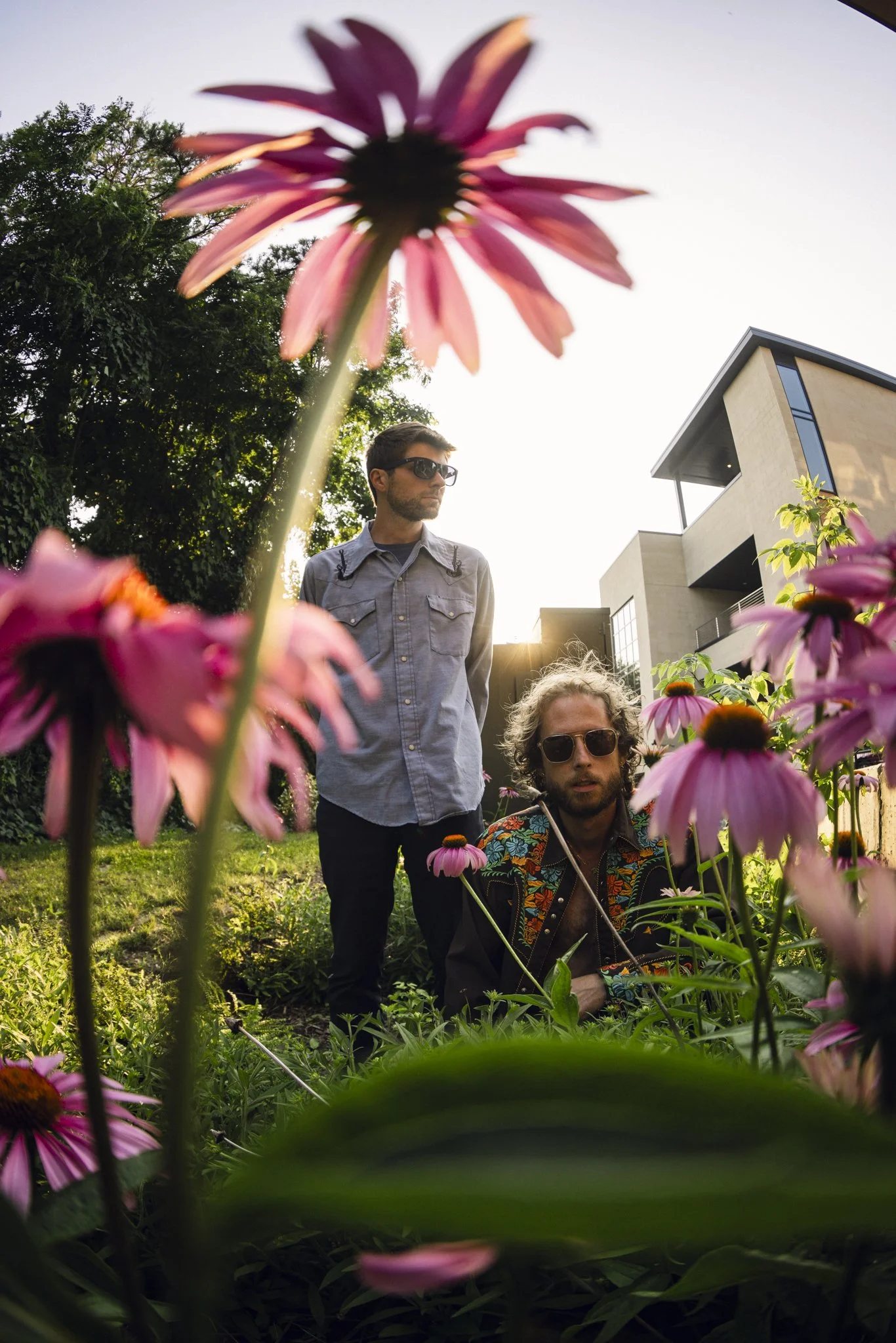 Two men in sunglasses are in a garden with pink flowers, with modern house architecture in the background during sunset.