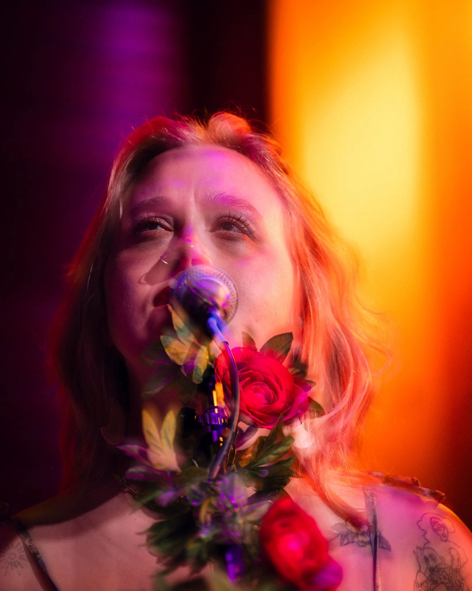 A woman with light skin singing into a microphone, with colorful stage lighting in pink, purple, orange, and yellow, and a large red rose near her face.