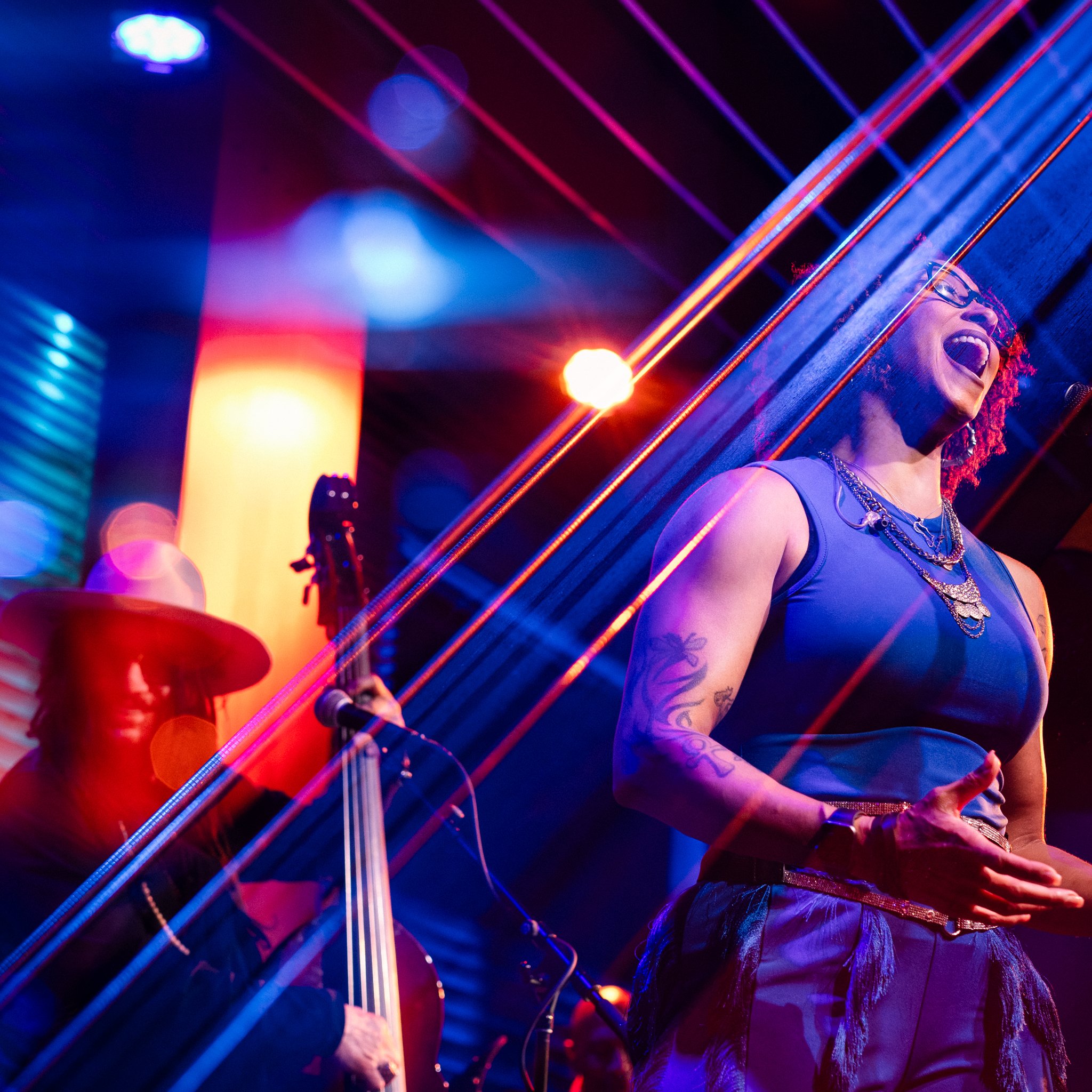 A woman singing on stage with a microphone, wearing a blue top and layered necklaces, with tattoos on her arms, during a live music performance with colored lights and a bassist in the background.
