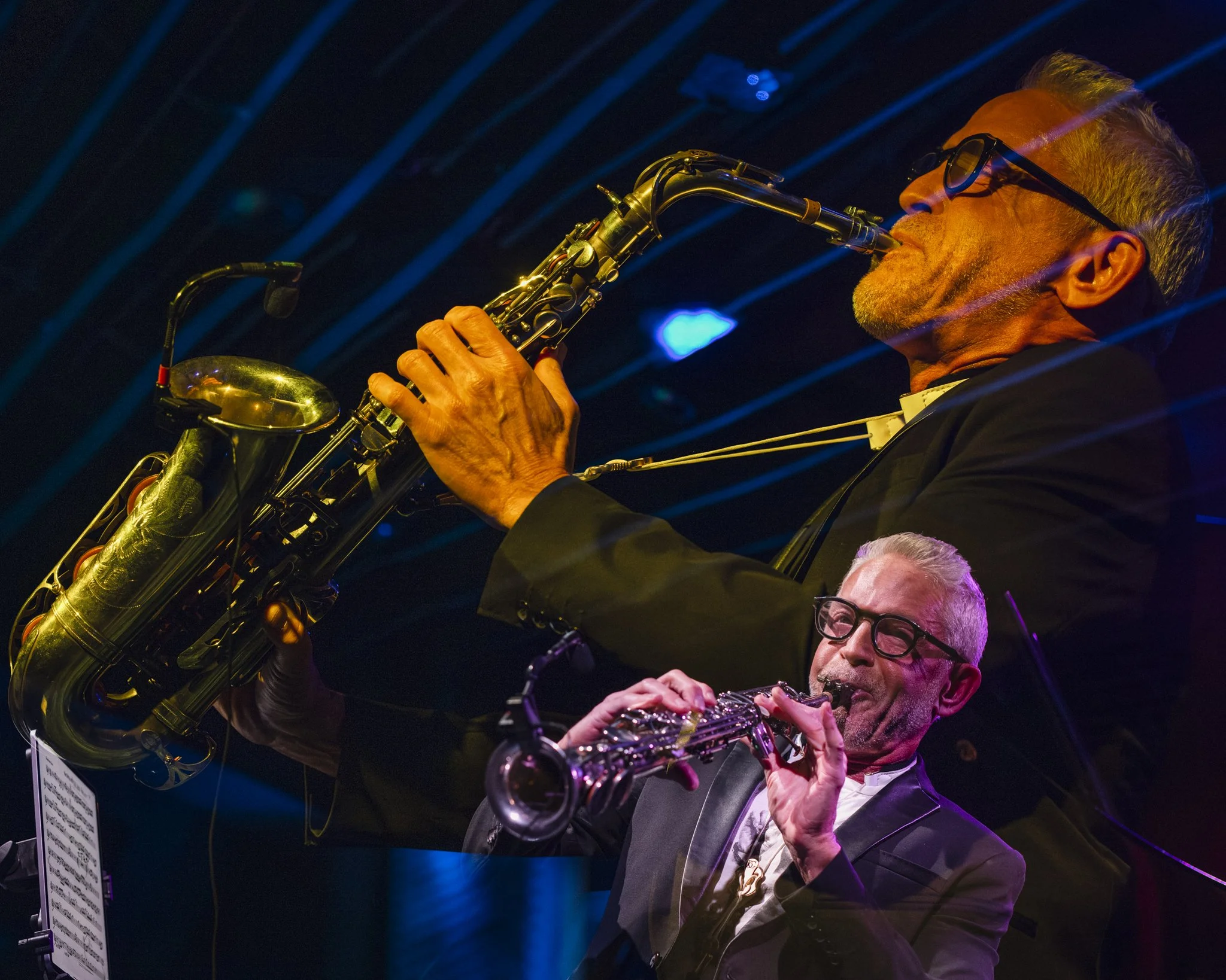 Two jazz musicians playing saxophone and clarinet on stage with colorful lighting reflections.