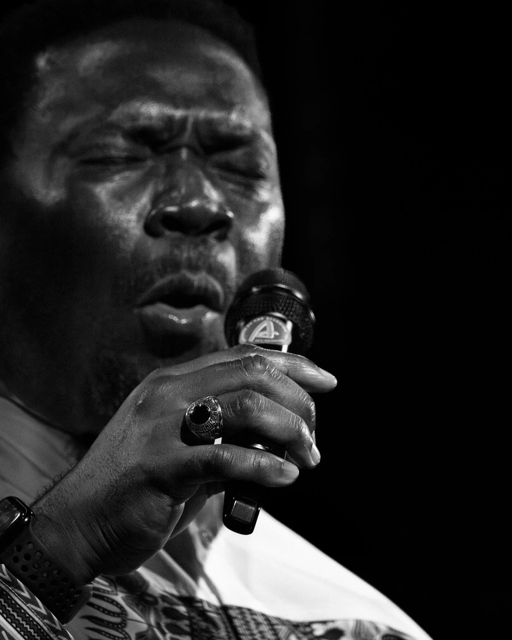 Black and white photo of a man singing into a microphone with his eyes closed, wearing a ring and a watch.