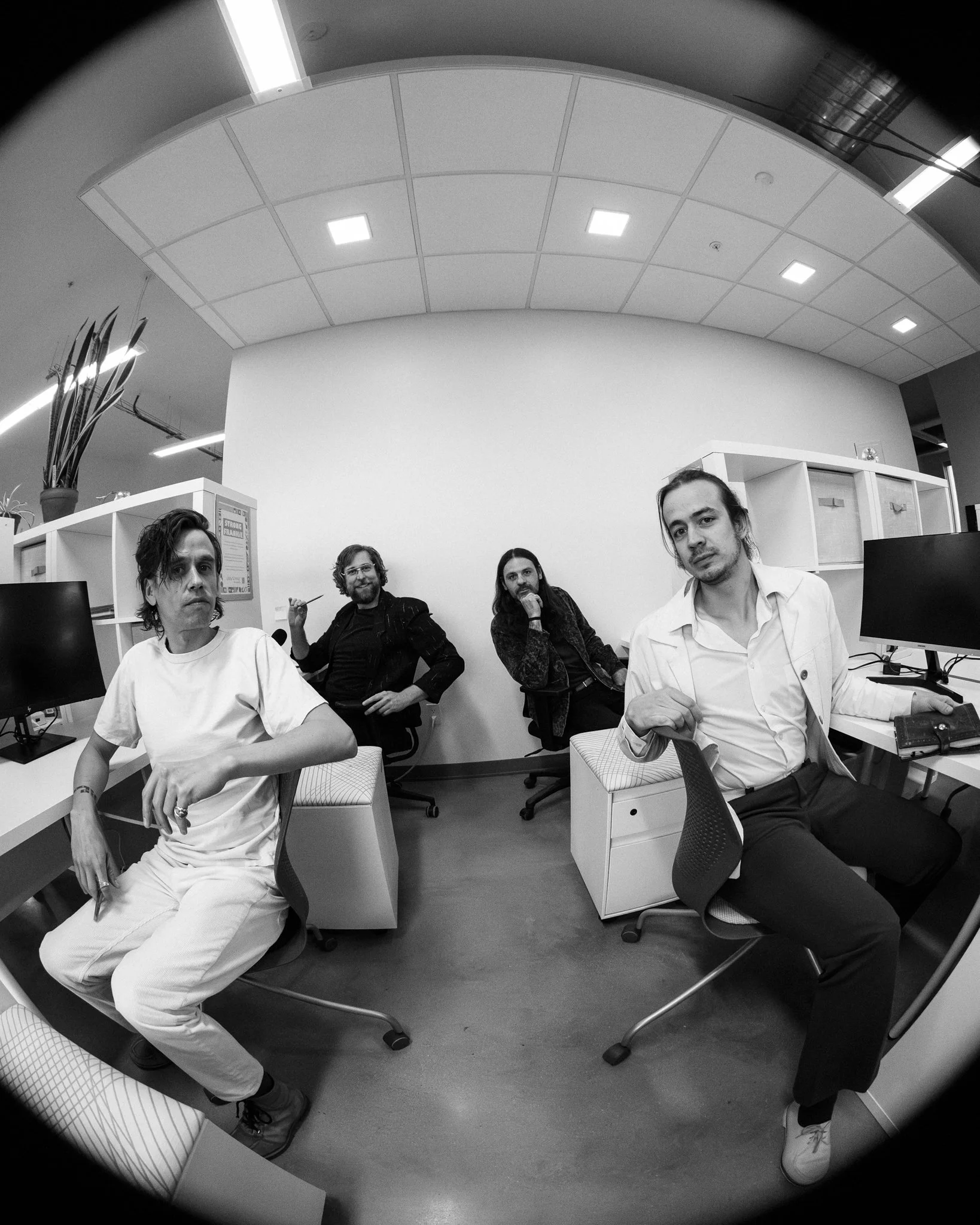 Four people in an office, sitting at desks with computers, looking at the camera. The photo is in black and white, and taken with a fisheye lens, creating a curved effect.