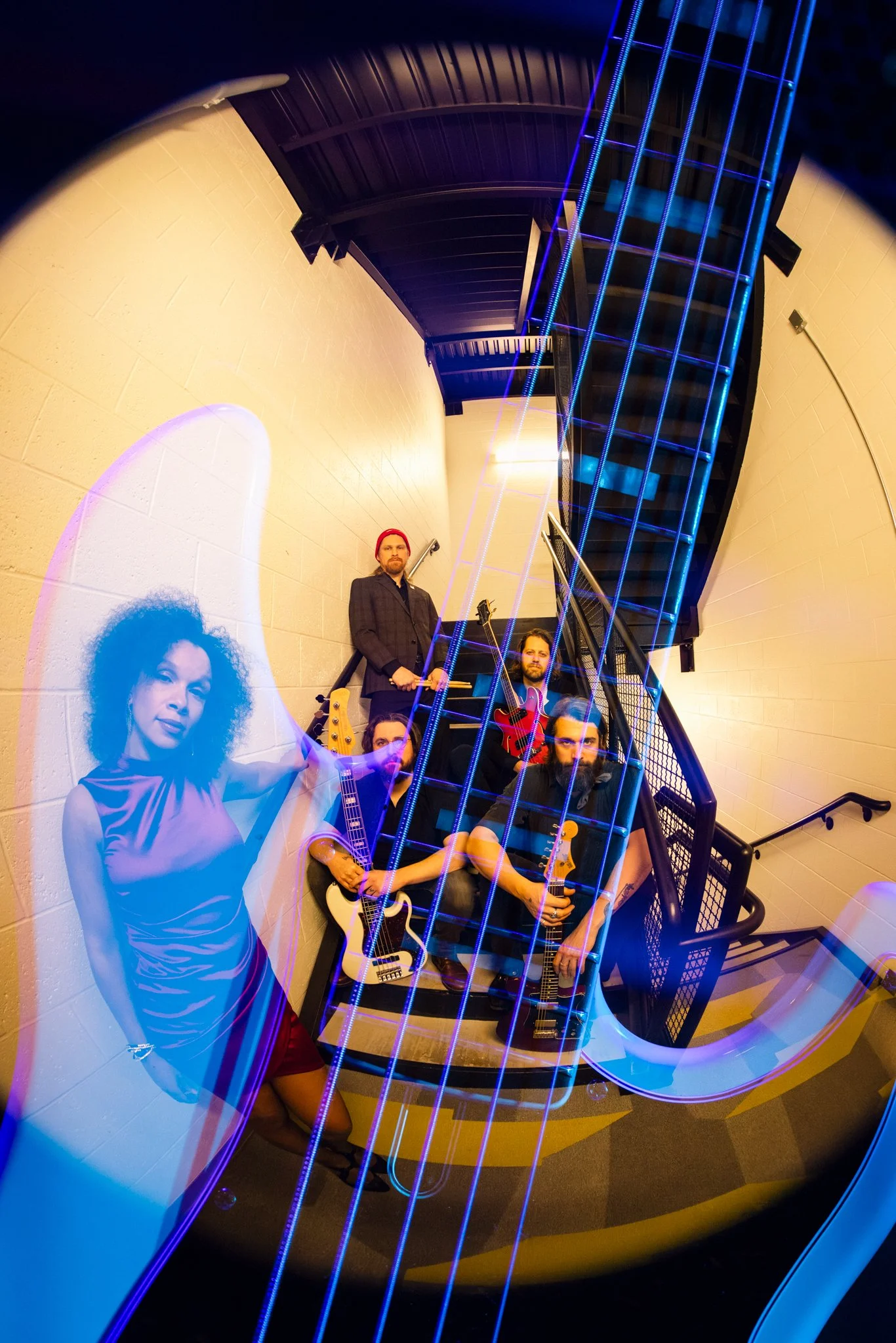 A laser-cut, fish-eye perspective photo of a band with two women and three men, some holding guitars, in a stairwell with neon lighting and a large portrait of a woman superimposed in the foreground.