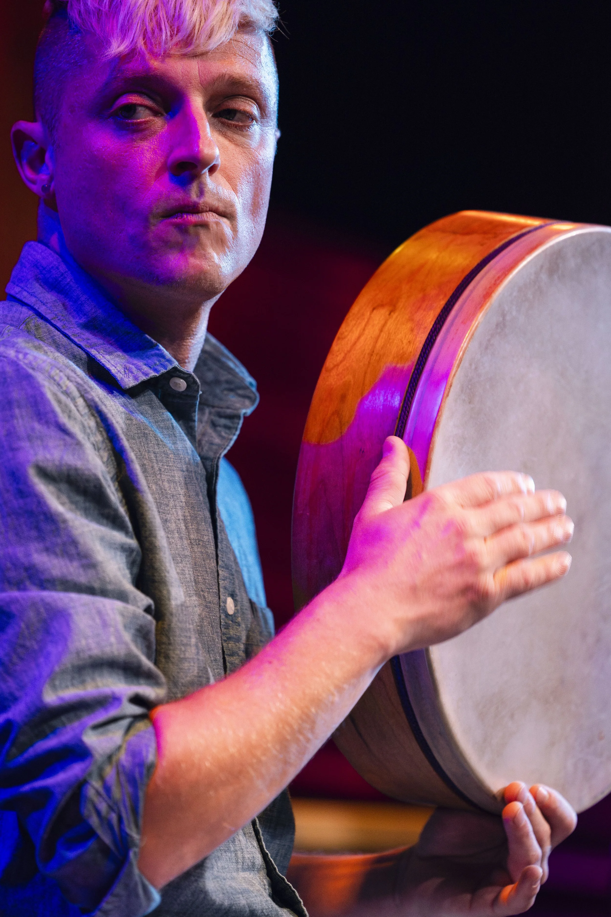 A person with short blonde hair, wearing a button-up shirt, holding a large drum with their right hand, in a performance setting with colorful stage lighting.