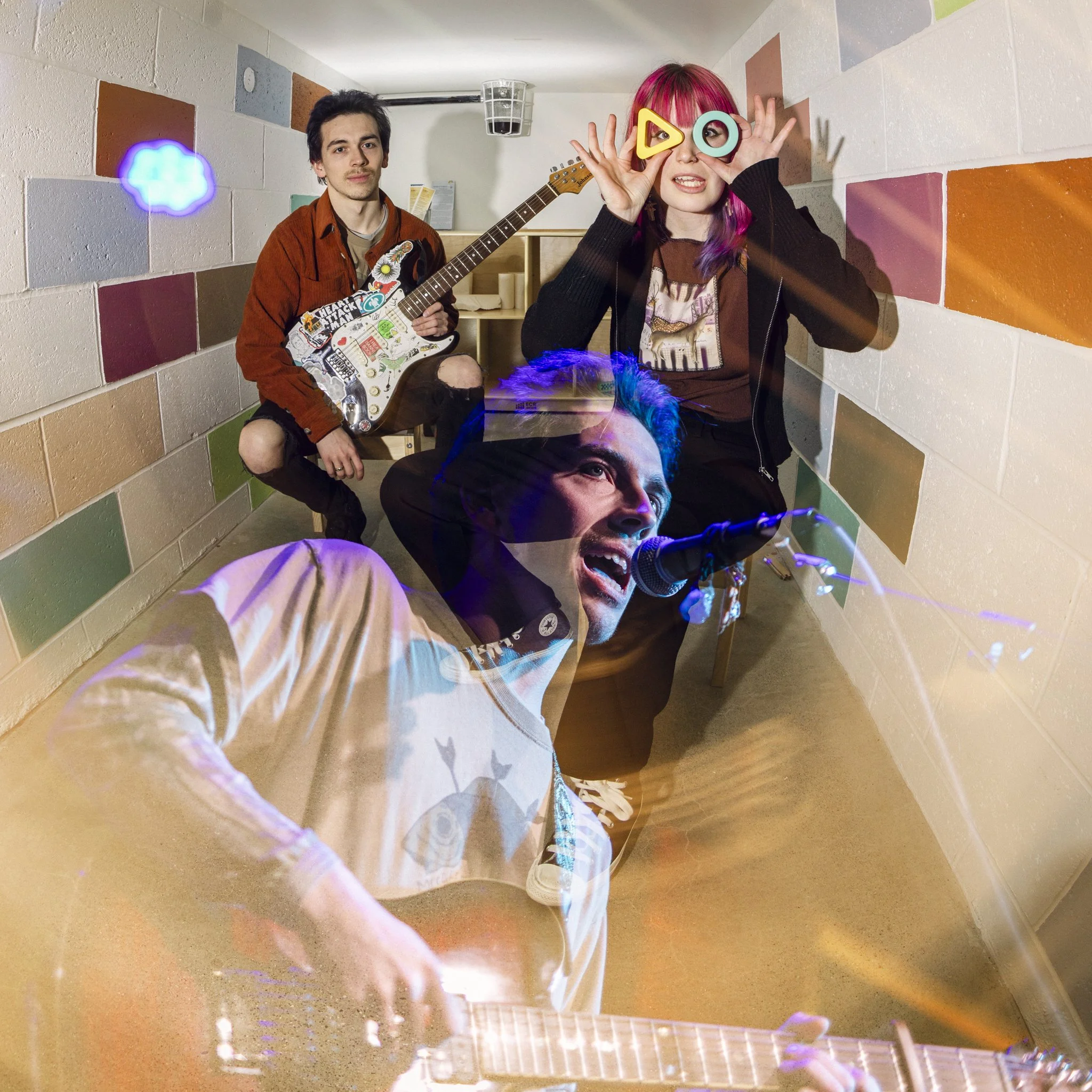 A colorful indoor scene with three young musicians, one playing guitar, one singing into a microphone, and one using funny glasses with geometric shapes, with a rainbow-colored wall in the background.